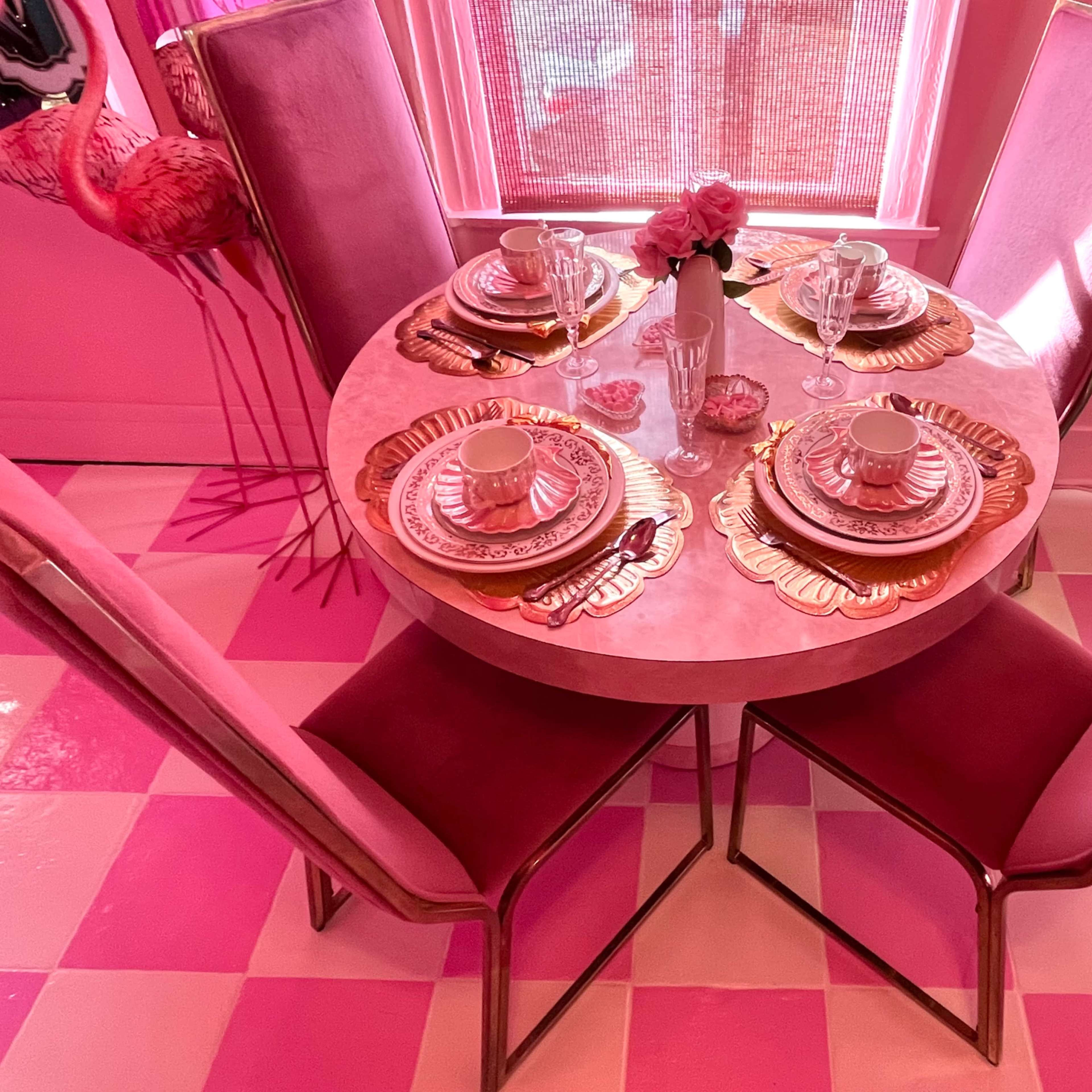 A round dining table is set with pink and gold plates, glassware, and a vase of flowers, surrounded by four upholstered chairs in a pink-themed room.