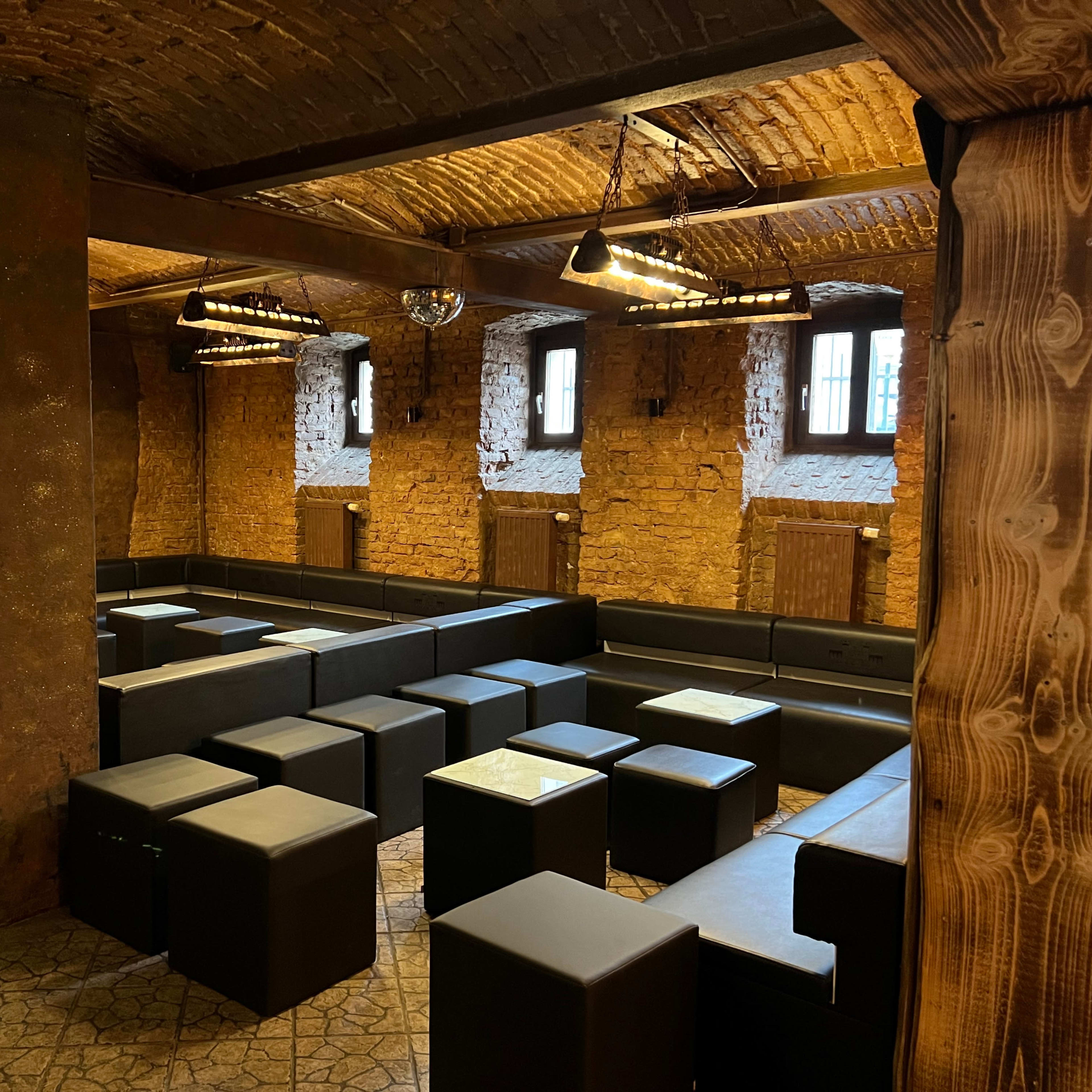 A dimly lit lounge area with black seating cubes arranged around small glass-topped tables against a rustic brick wall.