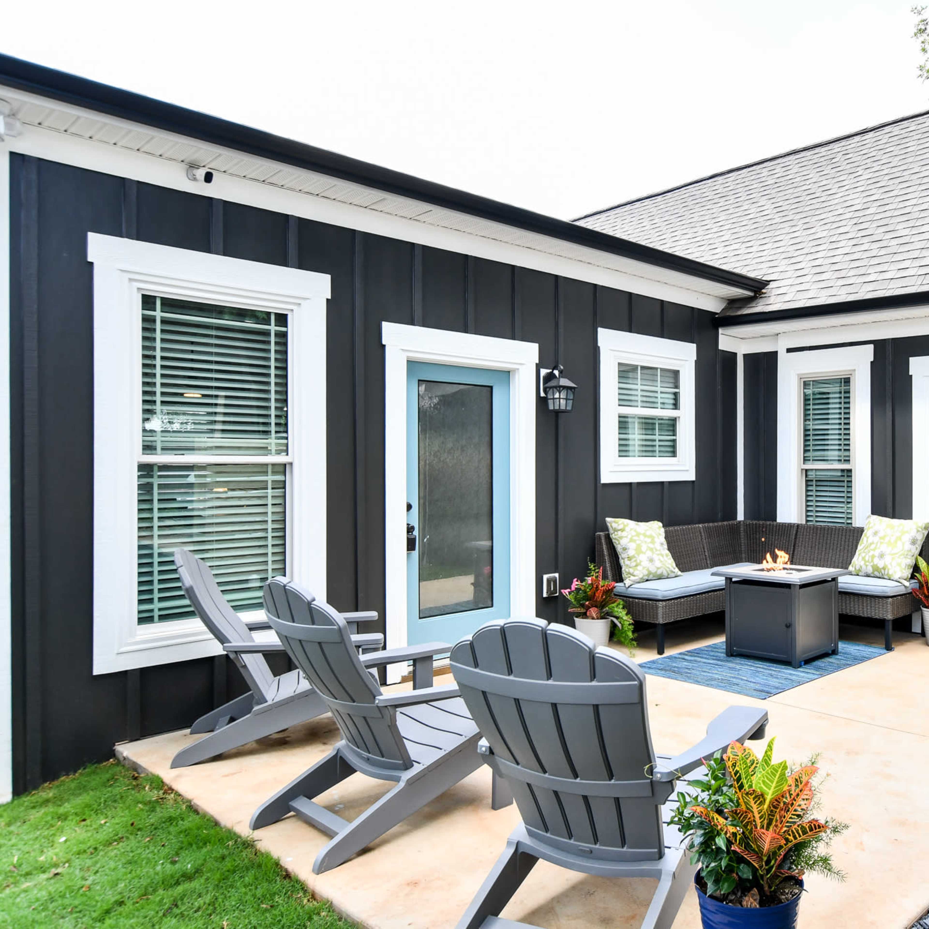 A modern outdoor patio with four gray Adirondack chairs, a fire pit, and a seating area surrounded by decorative plants.