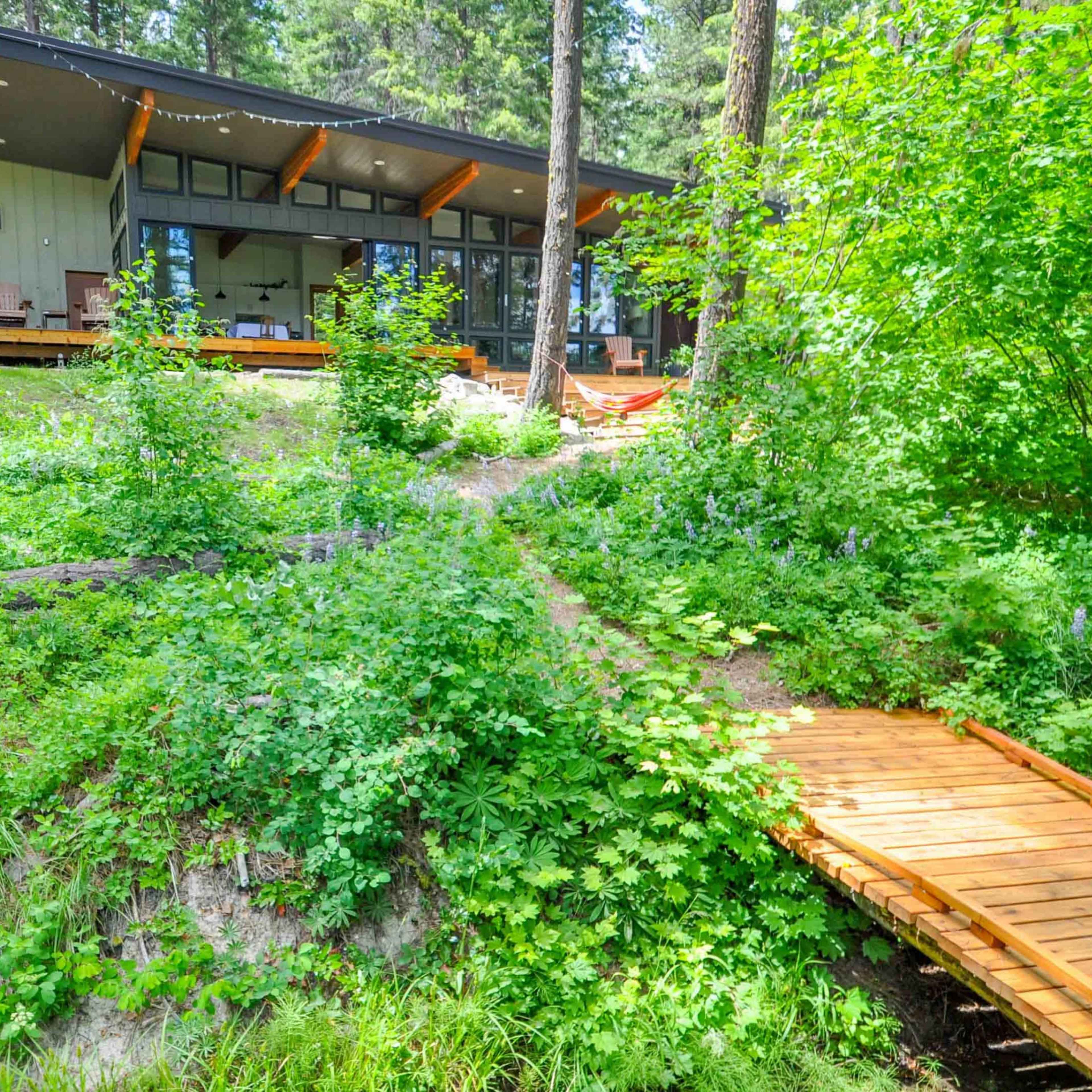 A wooden walkway leads to a modern house nestled among lush greenery and trees.