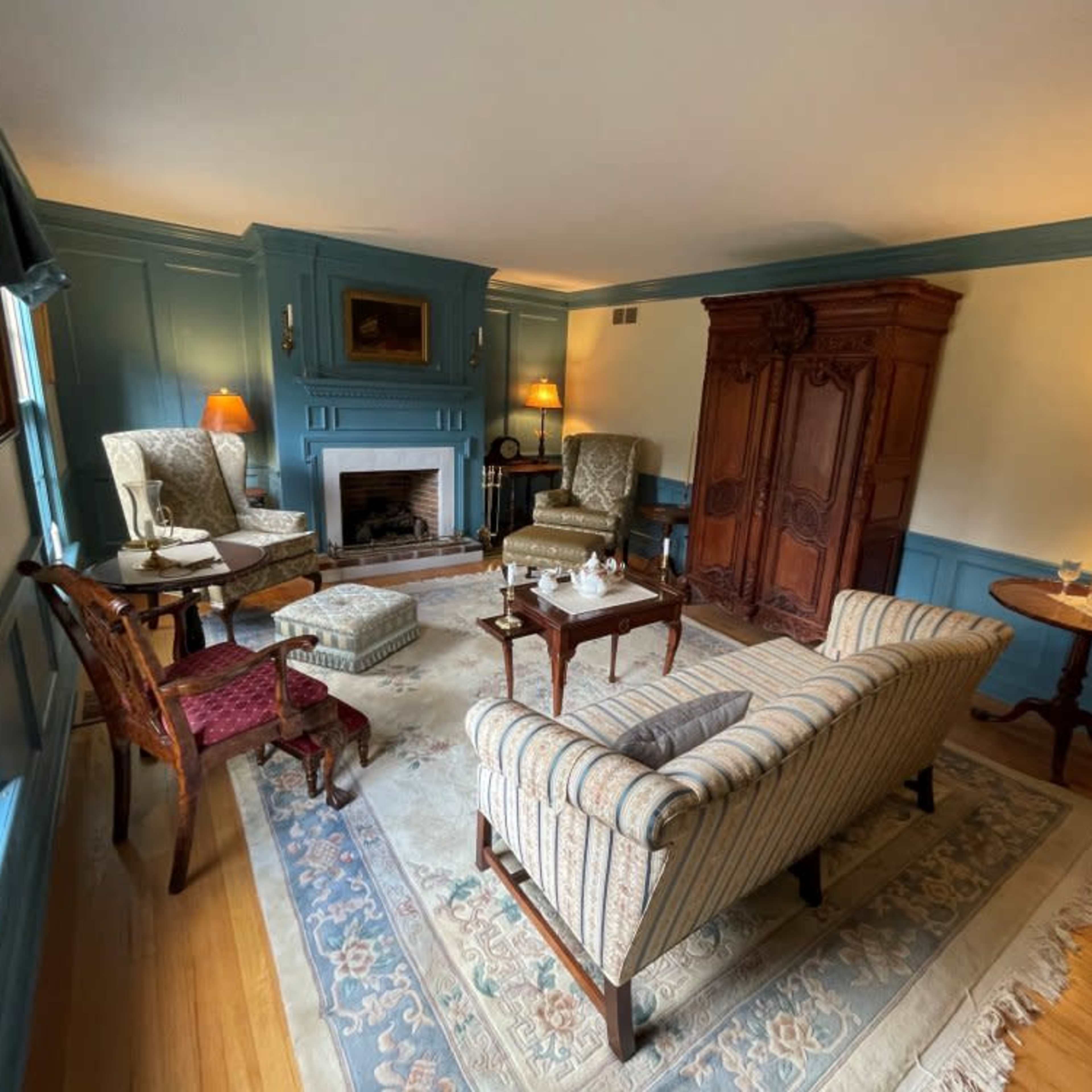 A living room with blue paneling, featuring a fireplace, two armchairs, a sofa, and a wooden cabinet, all arranged around a central area rug.