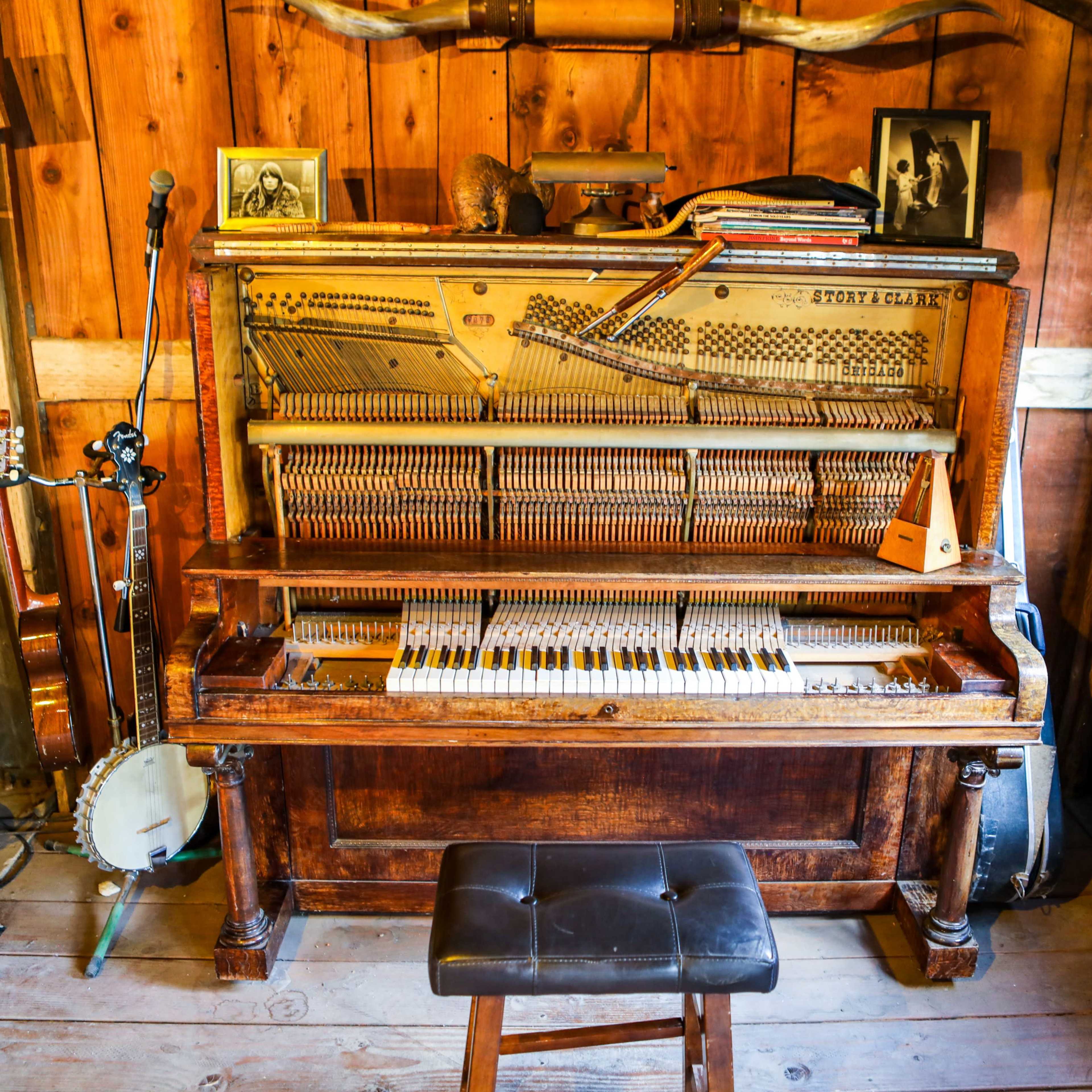An antique piano placed against a wooden wall, accompanied by a microphone, a banjo, and several photographs on the shelves above.
