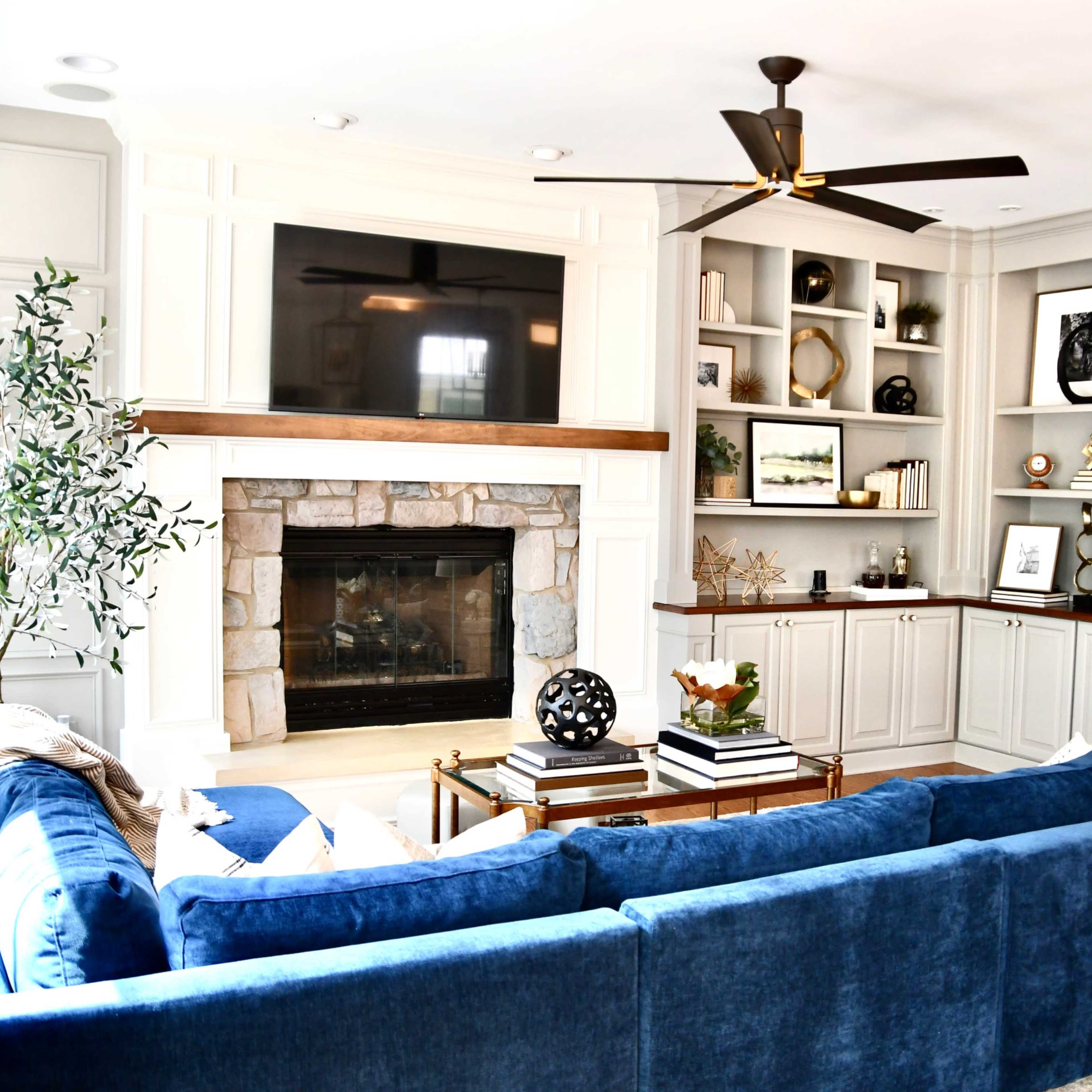 A spacious living room features a blue sectional sofa, a stone fireplace, built-in shelving with decorative items, and a ceiling fan.