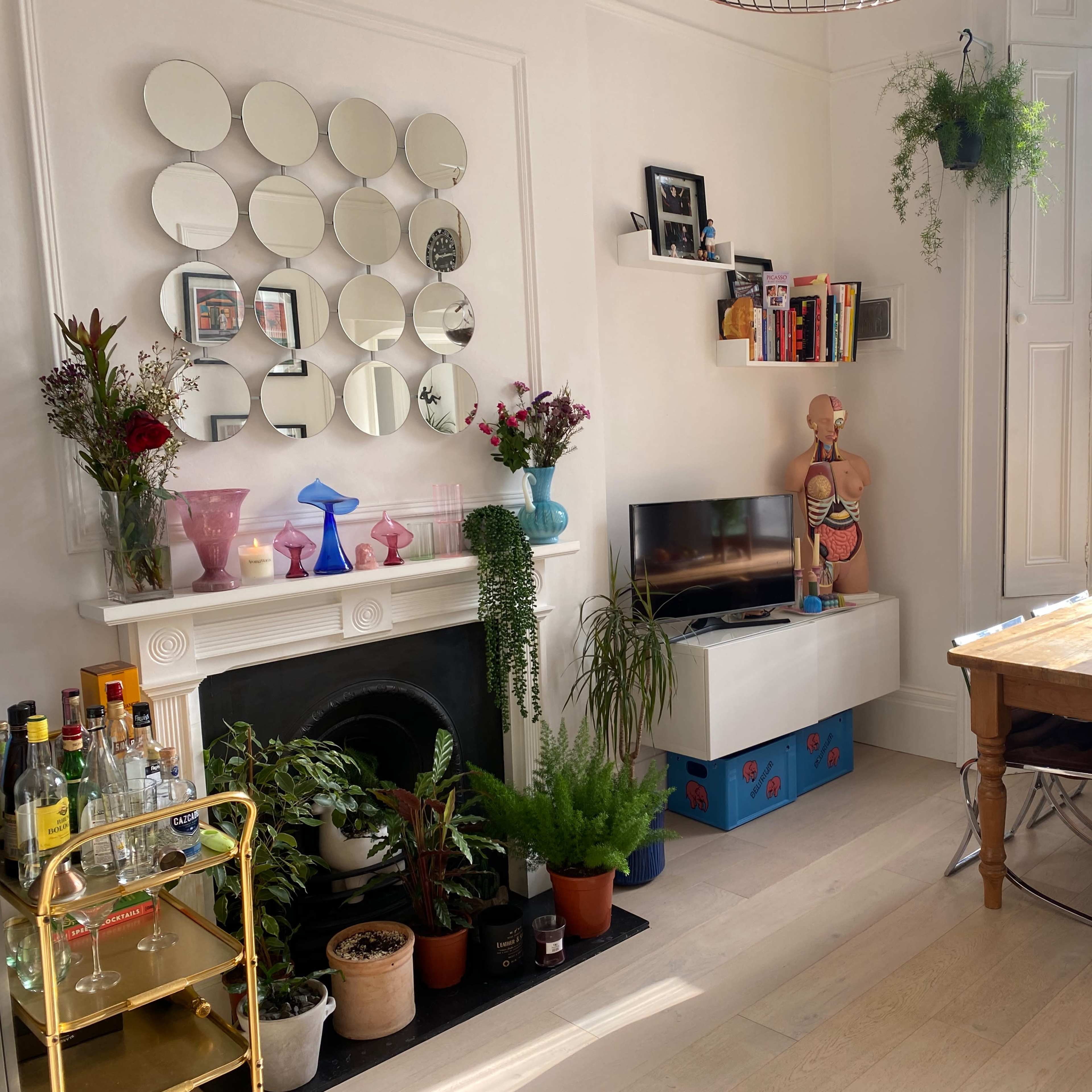 A stylish living room with a large window, a decorative mirror above a fireplace, a TV, a dining table, and various plants and artworks.