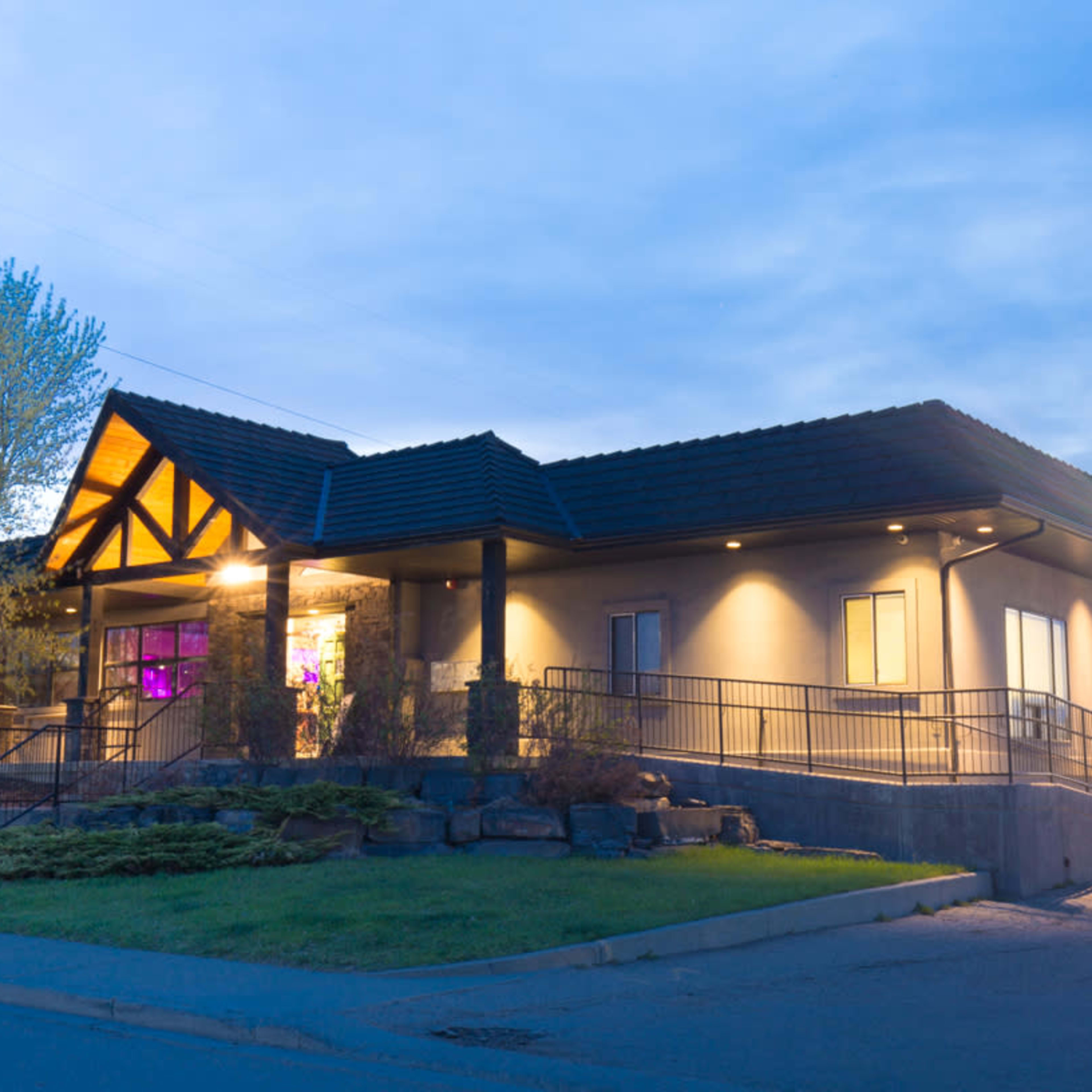 A modern building with a sloped roof, a ramp leading to the entrance, and illuminated windows under a twilight sky.