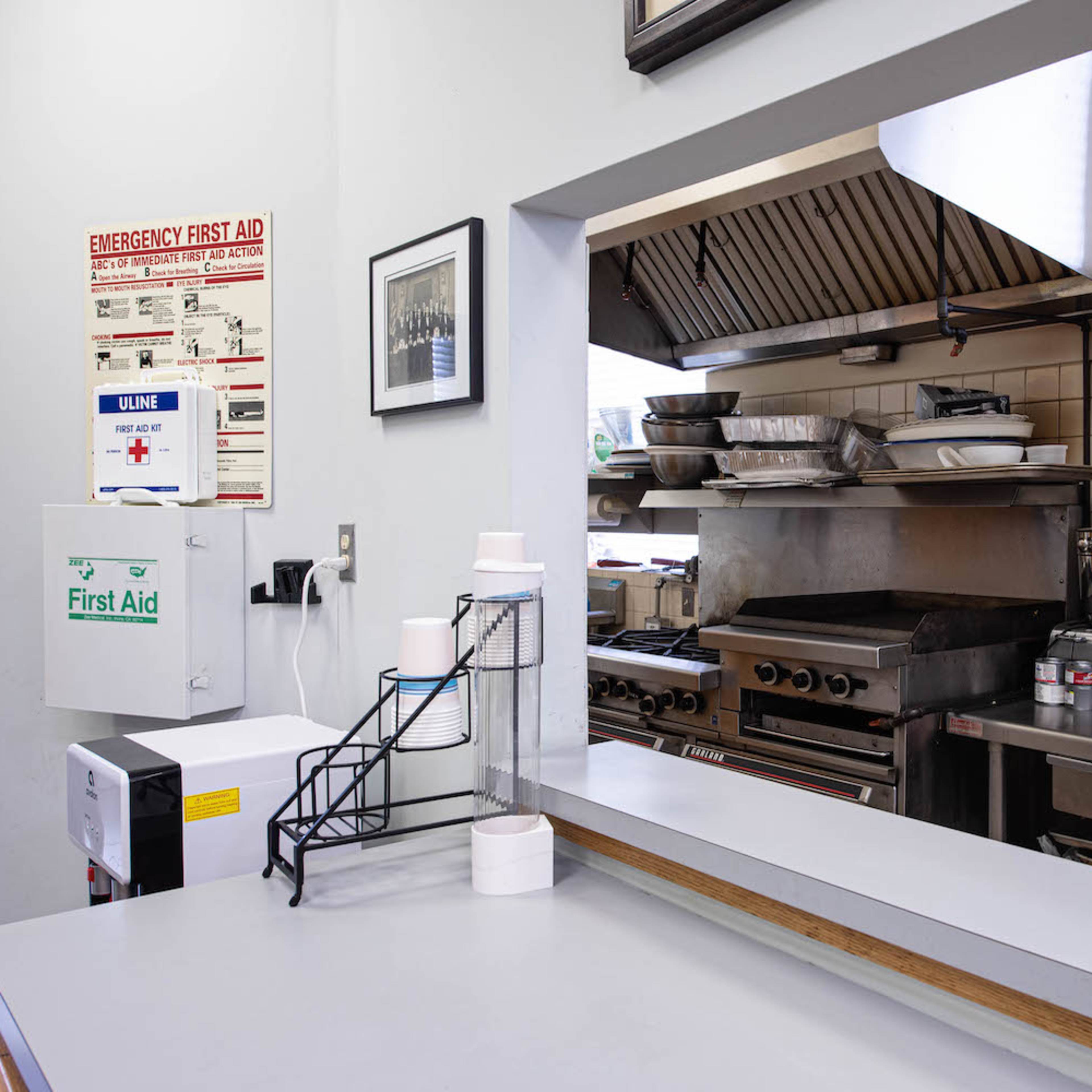 A commercial kitchen with various appliances, including a stove and a ventilation hood, and a counter area equipped with a first aid station.