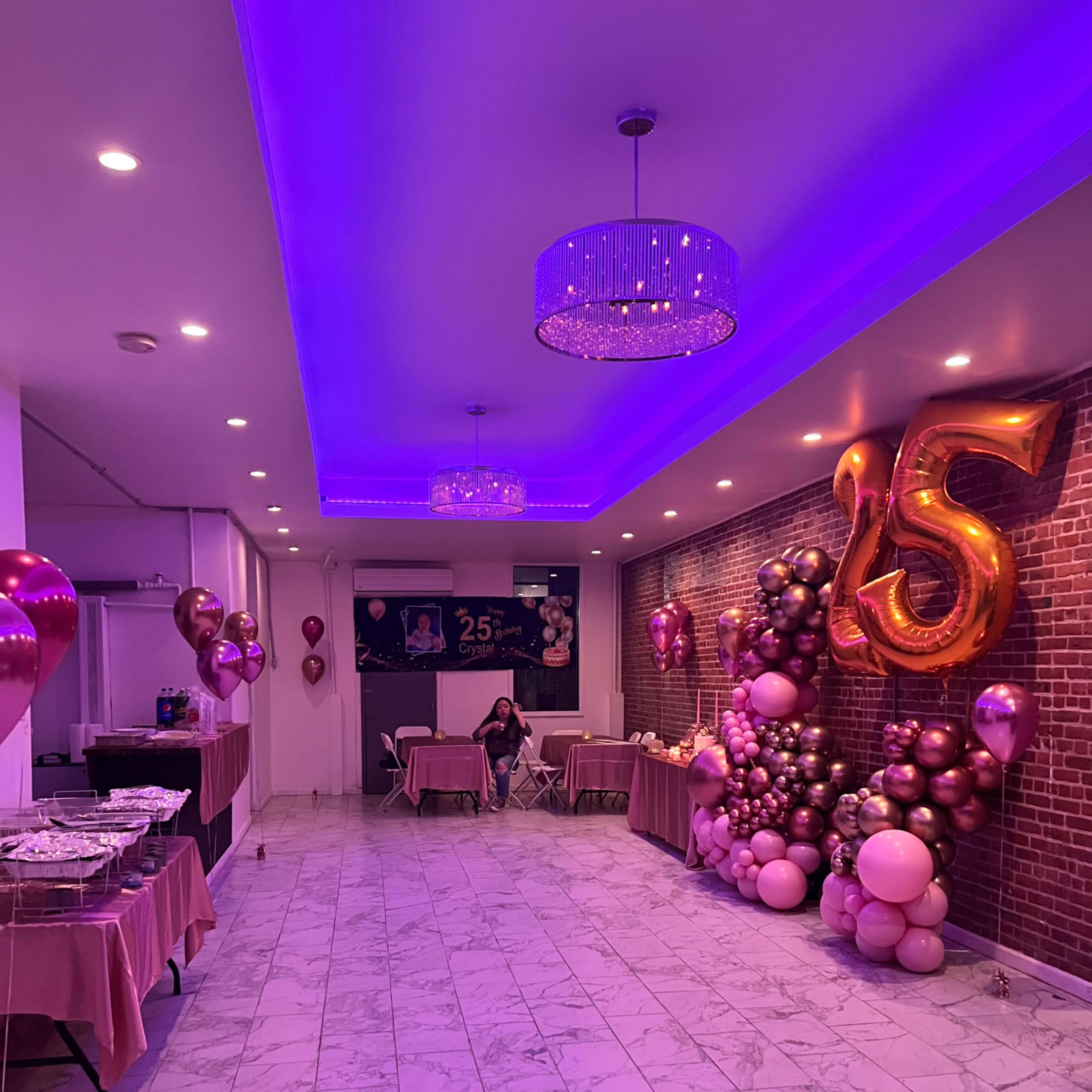 A festive event space decorated with balloons and a large "25" display, featuring tables set up along the walls and a vibrant purple light overhead.