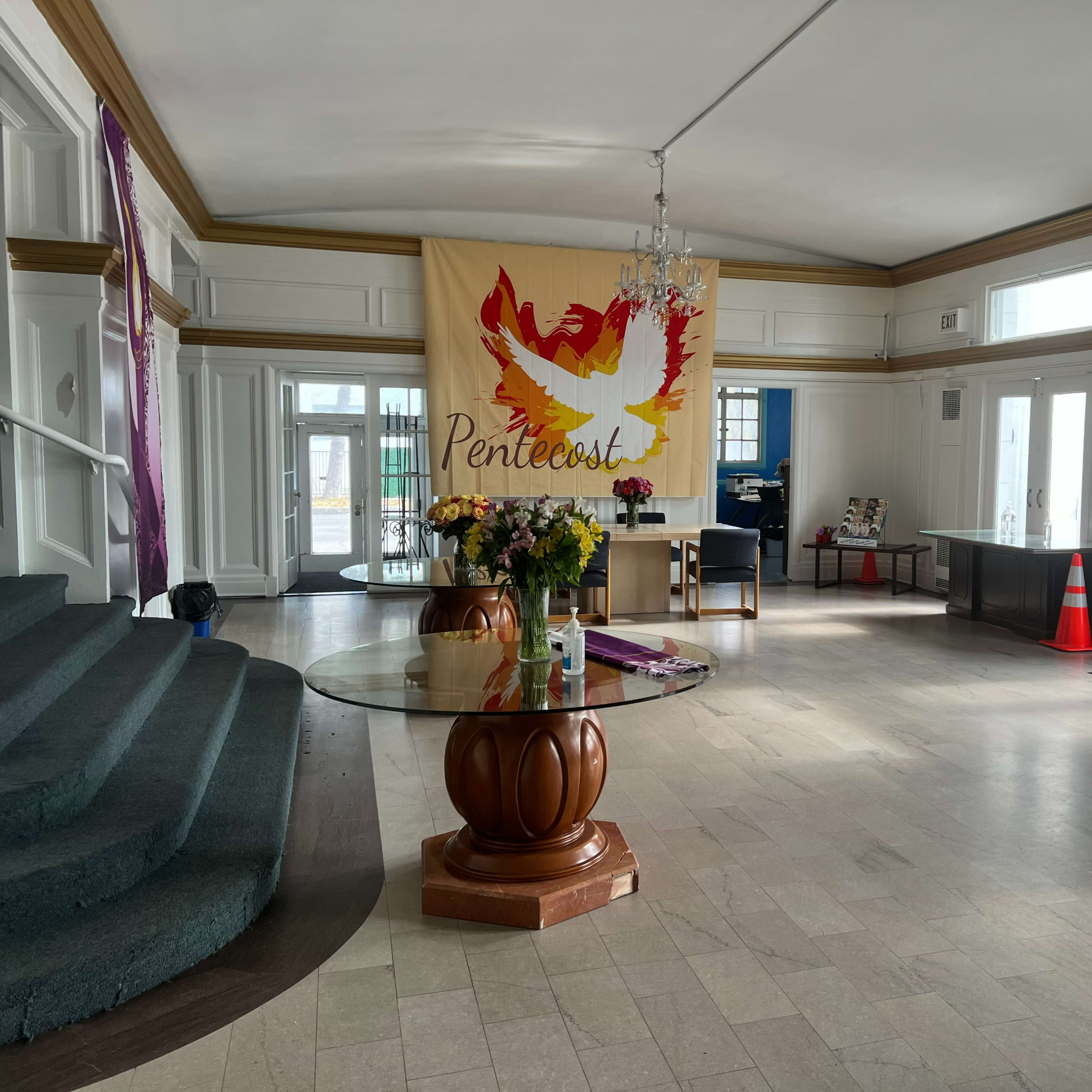 A spacious lobby with a circular glass table, a large banner reading "Pentecost," and several chairs near the entrance.