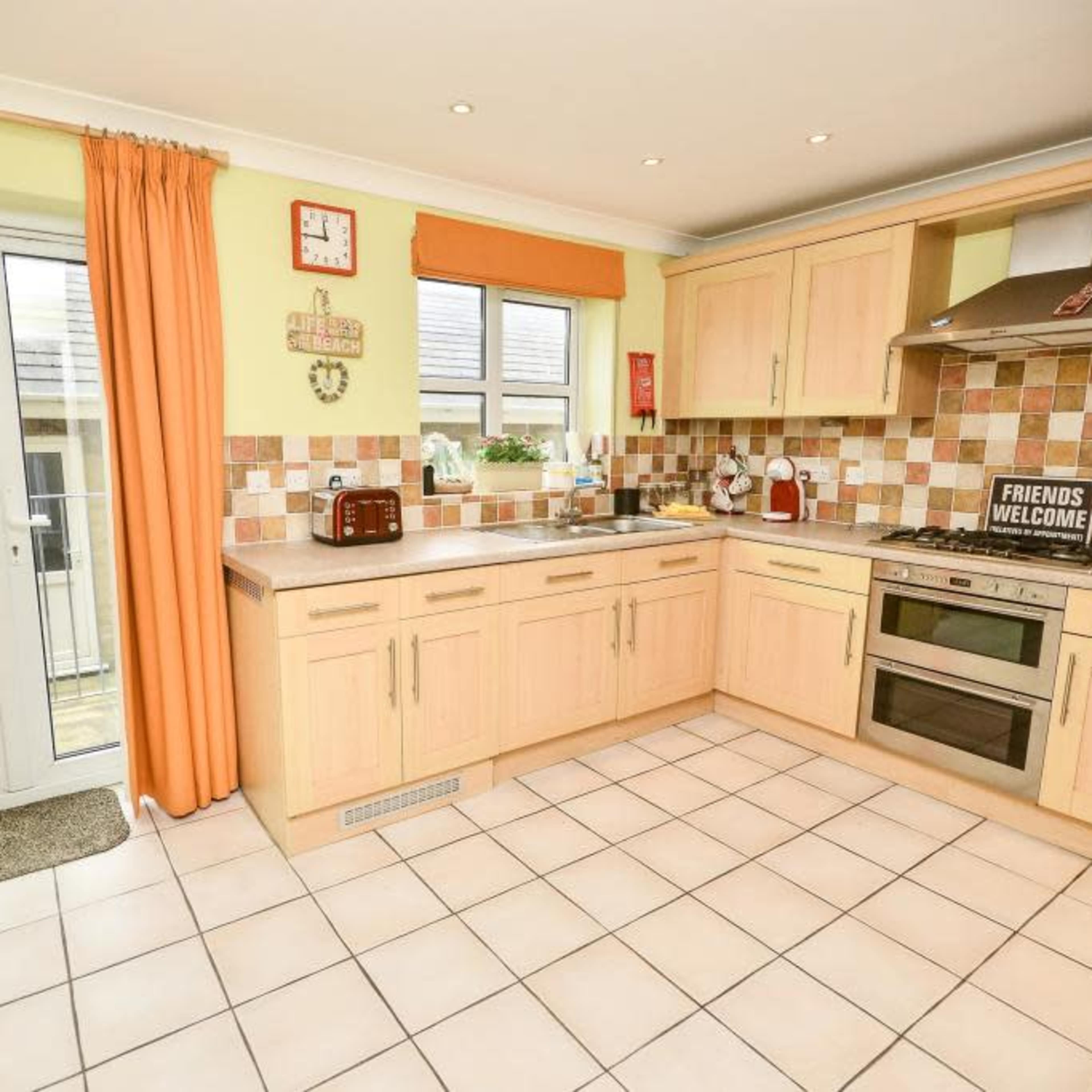 A modern kitchen with light wooden cabinetry, tiled flooring, and large windows allowing natural light.