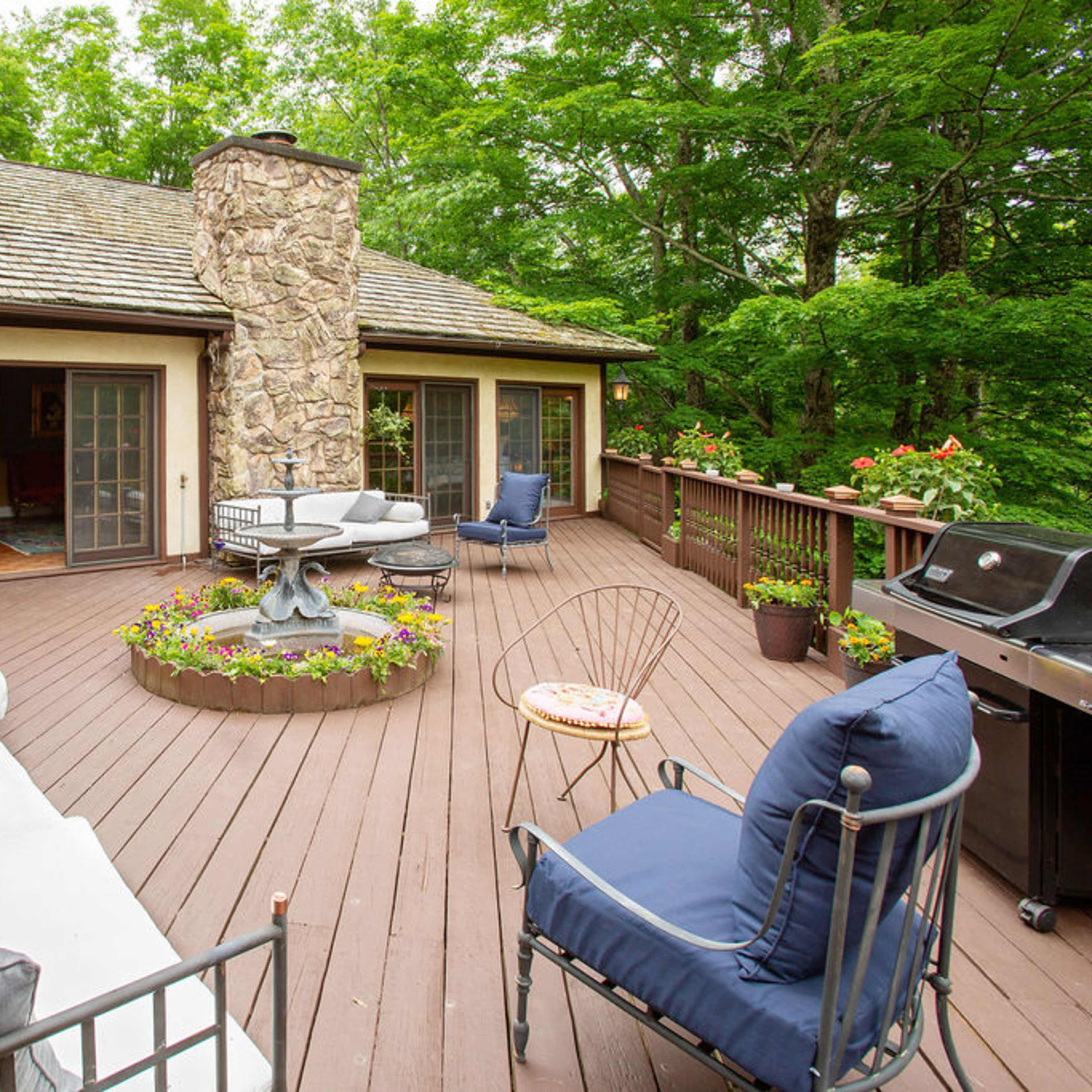A wooden deck features a stone chimney, seating areas, and a circular flowerbed surrounded by lush greenery.