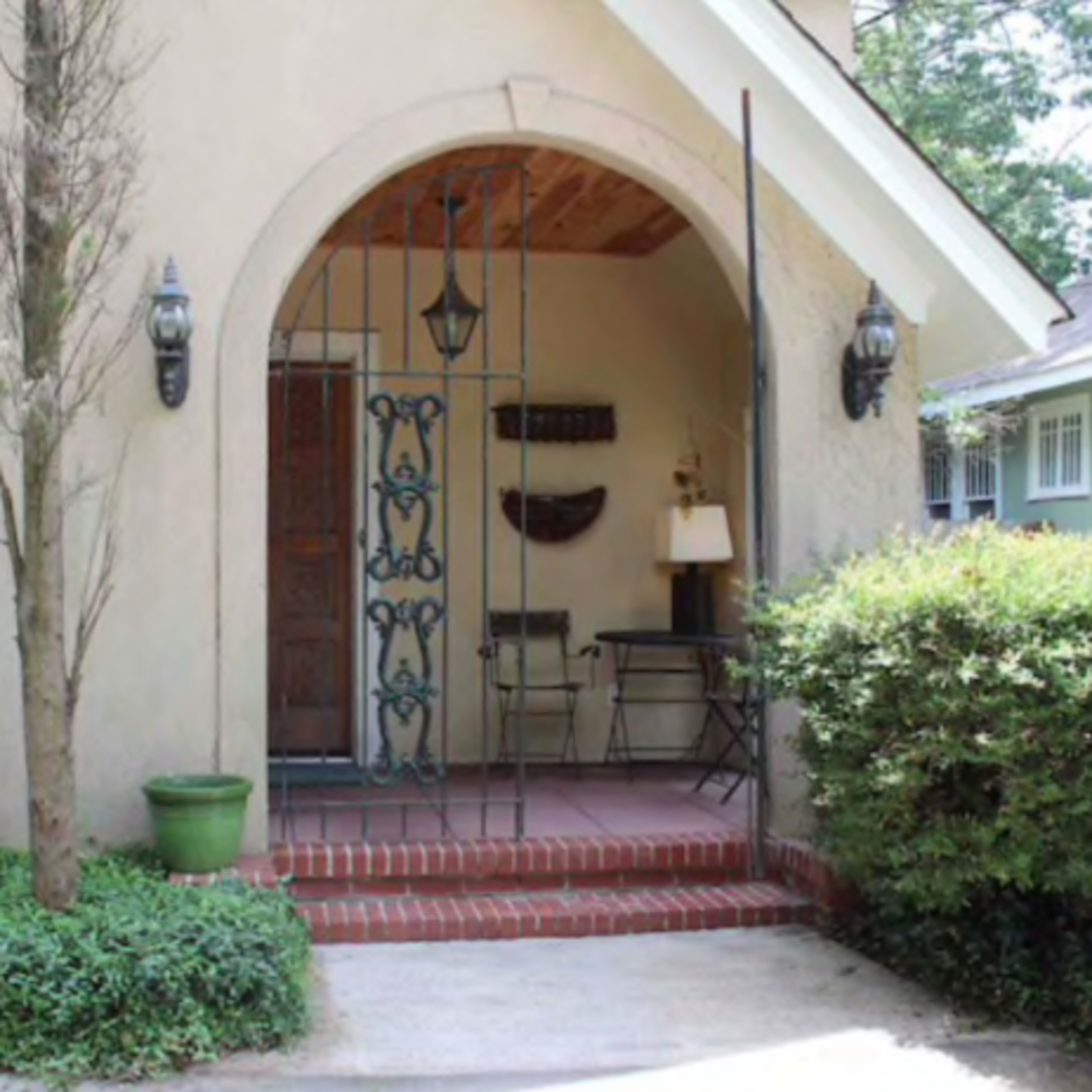 A small entrance with an arched doorway, featuring a wrought iron gate, brick steps, potted plants, and a table with a lamp inside.