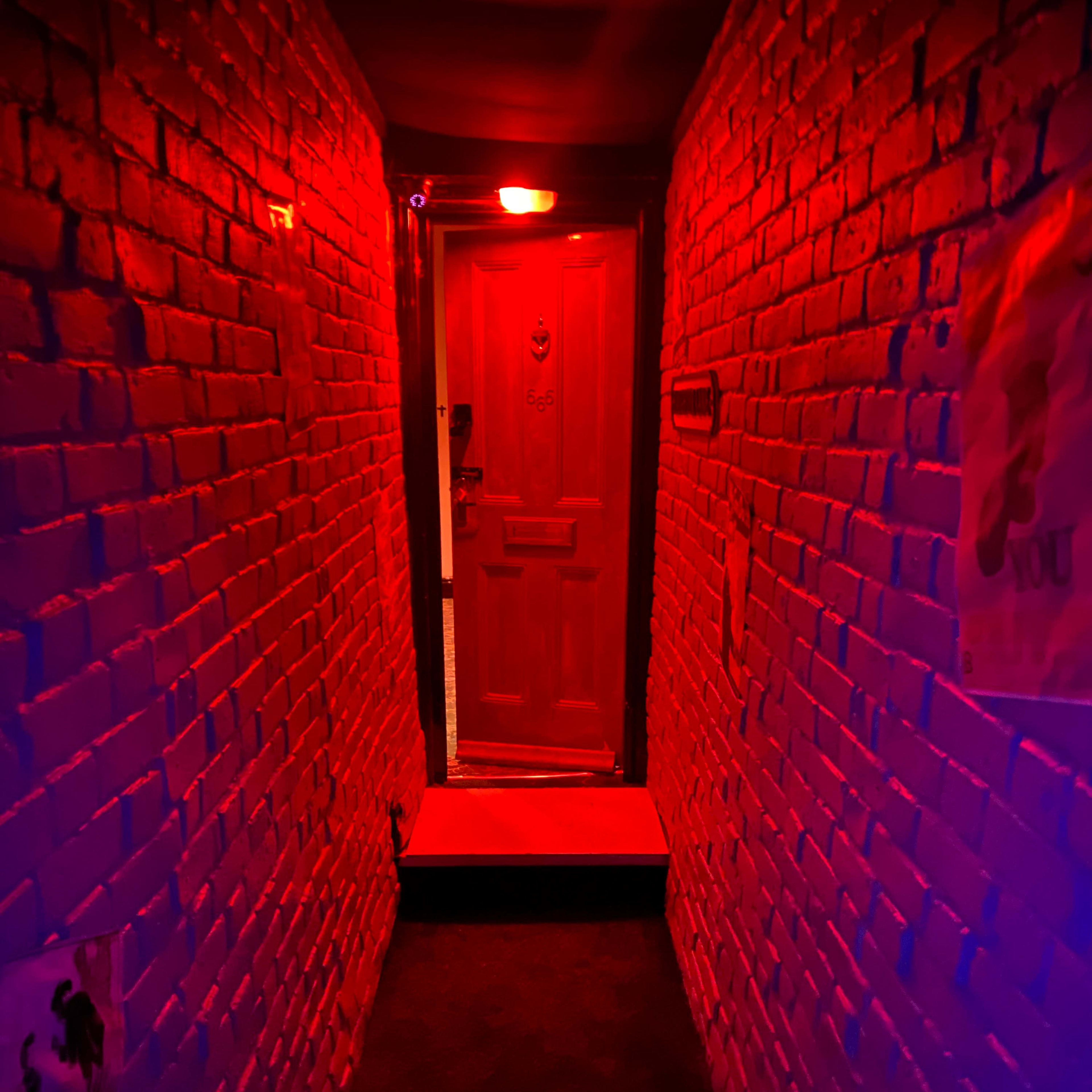 A narrow corridor with red-lit brick walls leads to a wooden door at the far end.