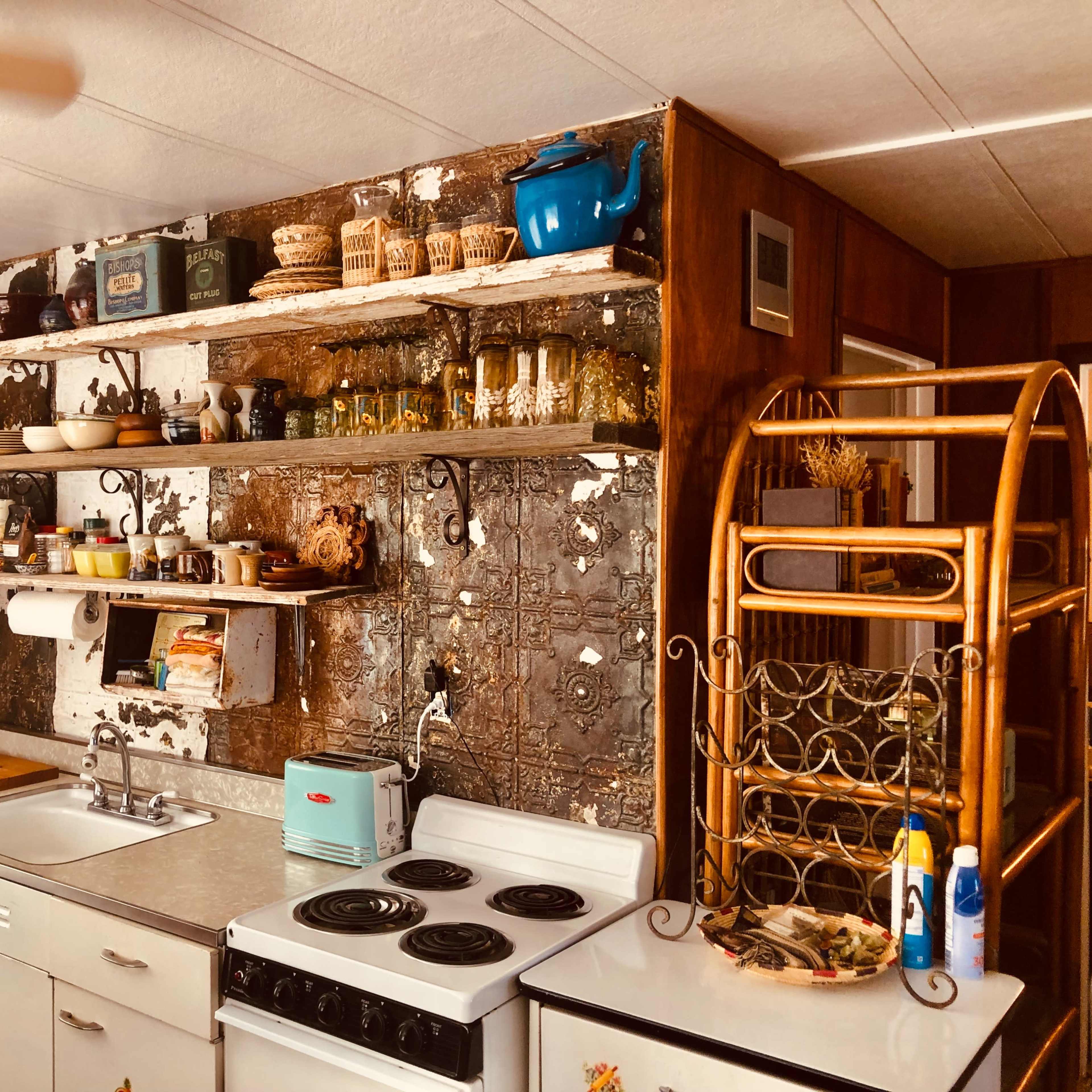 A kitchen with a sink, stove, and shelves displaying various dishes and decorative items against a wall featuring a mix of wood and metal.