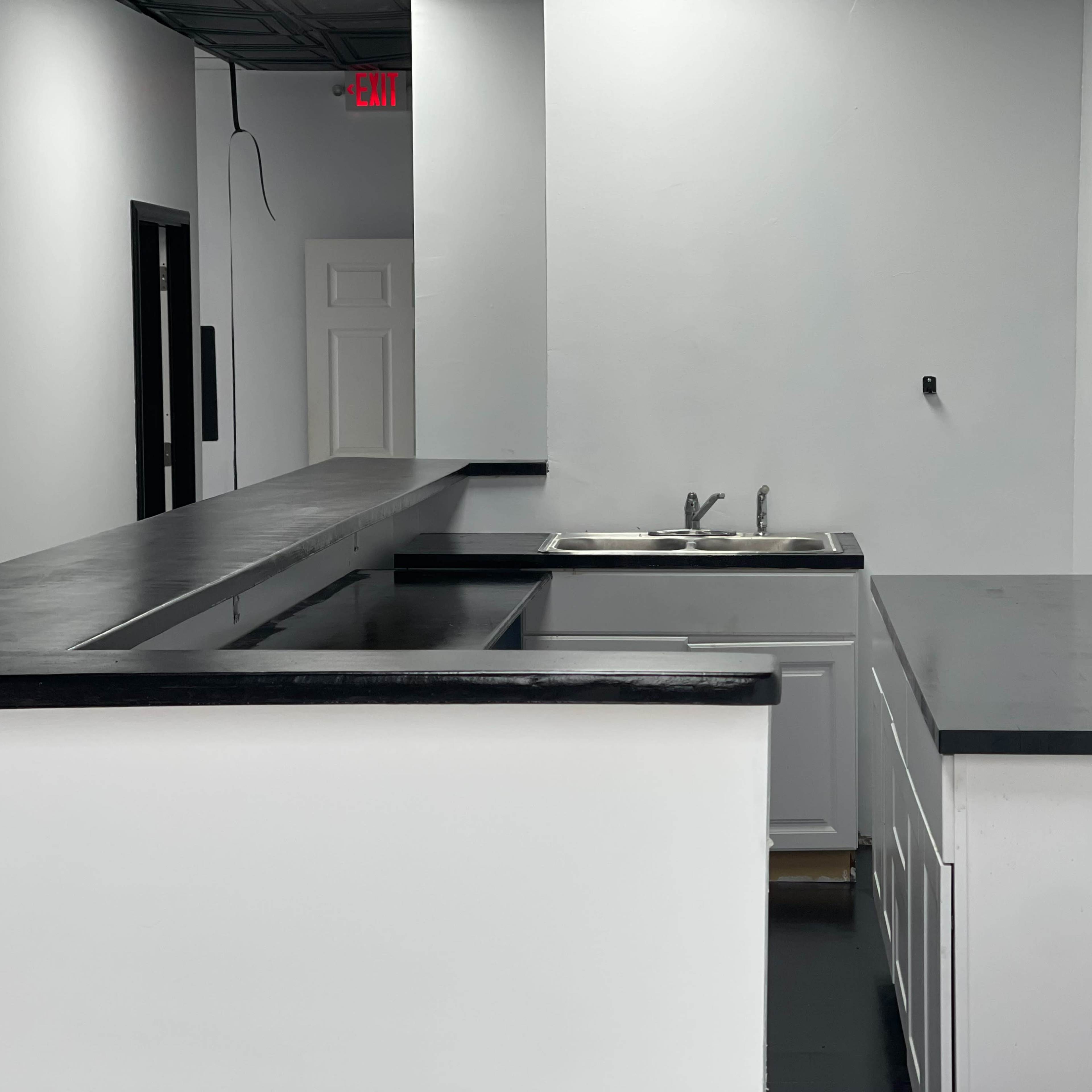 A minimalist kitchen area with a black countertop, a sink, and white cabinetry against a plain wall.
