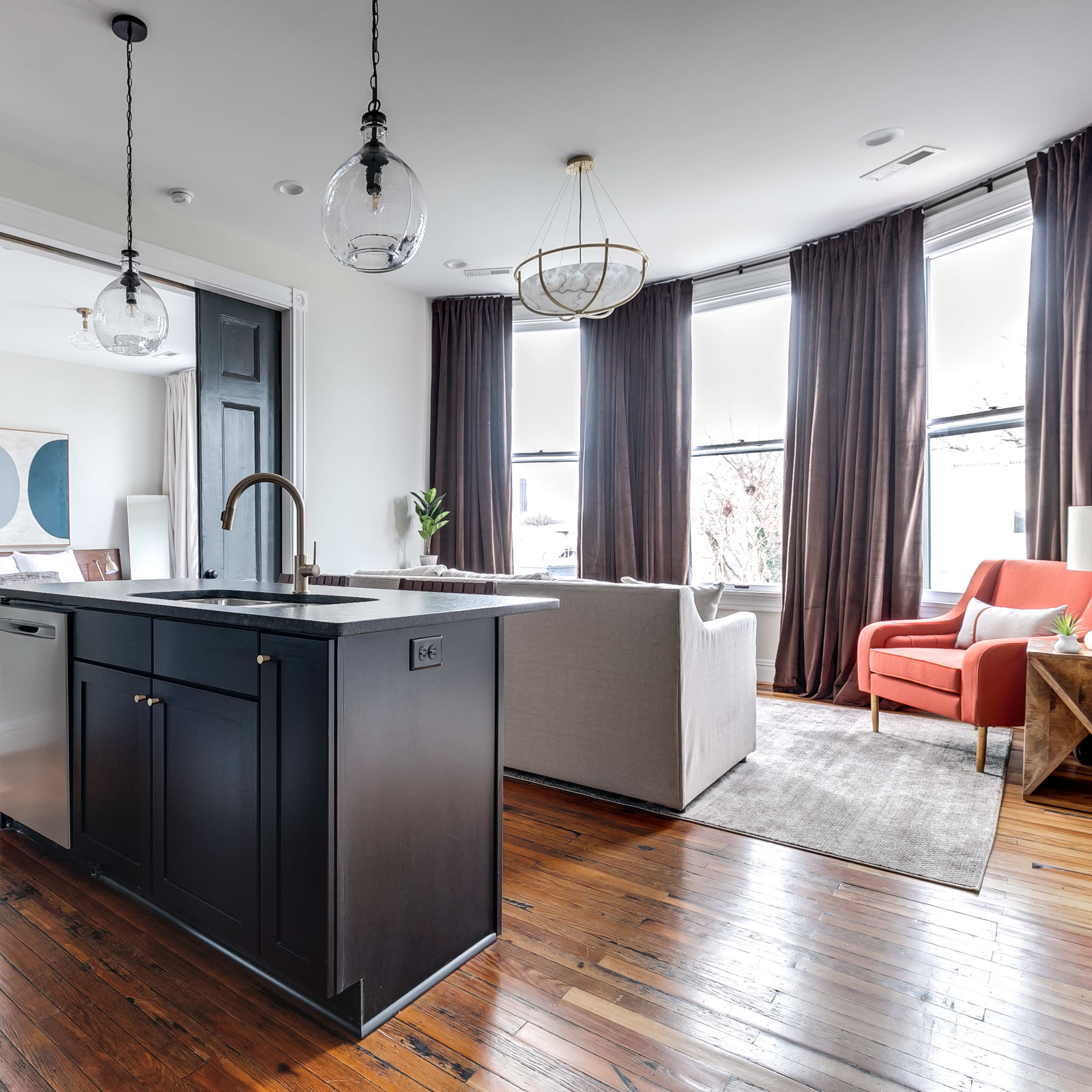 A modern kitchen features a central island with a dark finish, leading to an open living area with large windows and stylish furnishings.