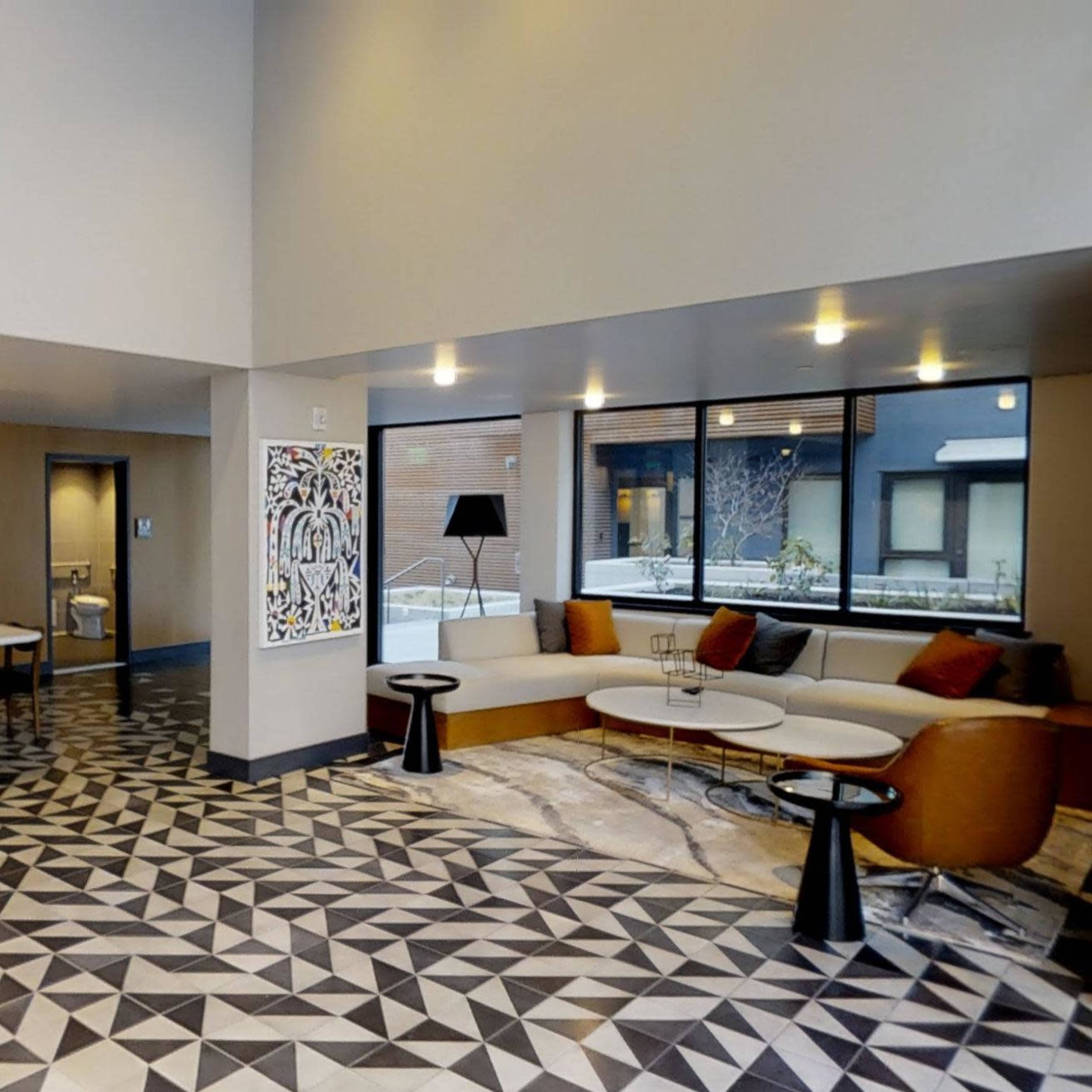 A modern hotel lobby featuring a patterned floor, a seating area with orange chairs, and a long table with hanging pendant lights.