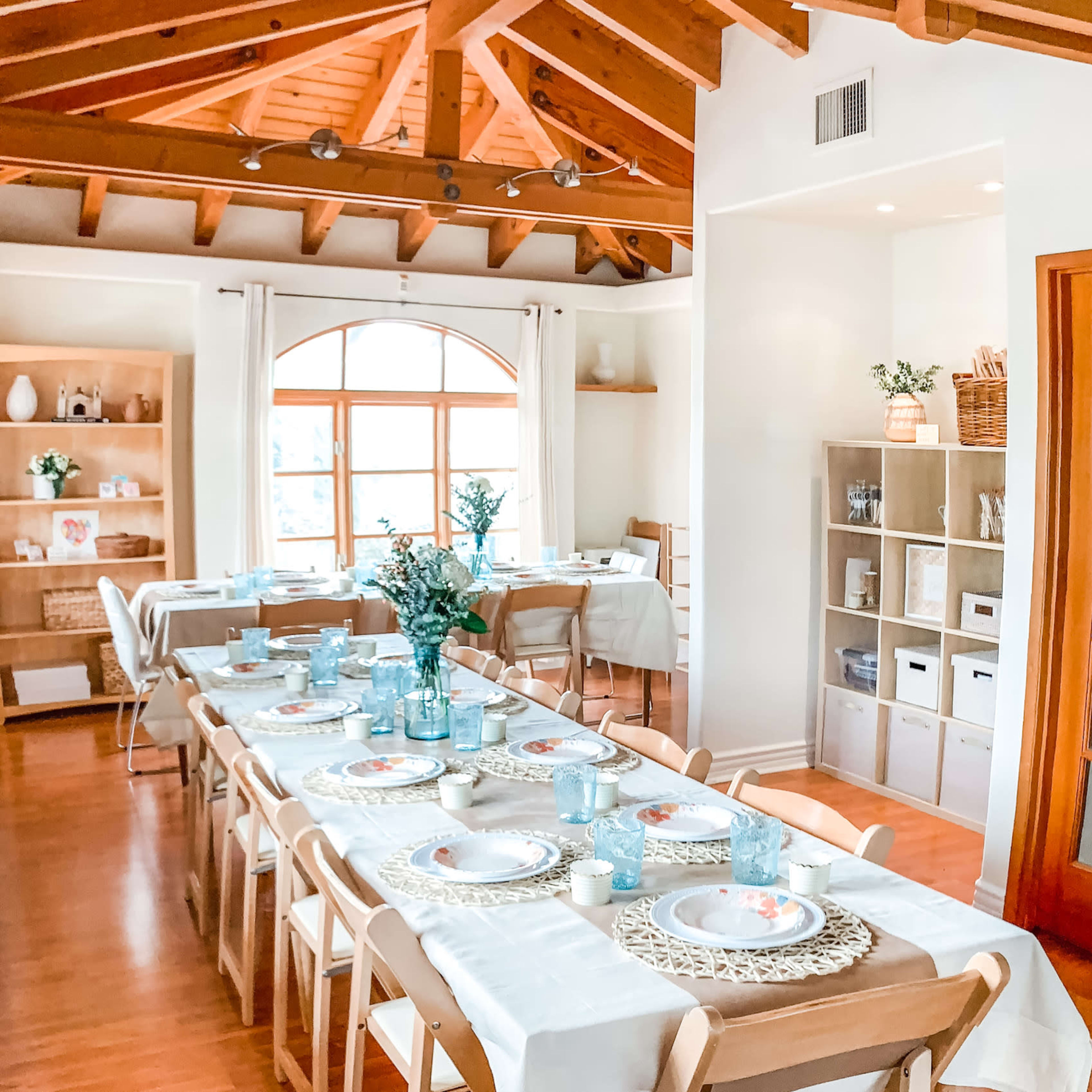 A bright dining area with wooden beams, a long table set with plates and glassware, and an adjacent shelving unit filled with decorative items.
