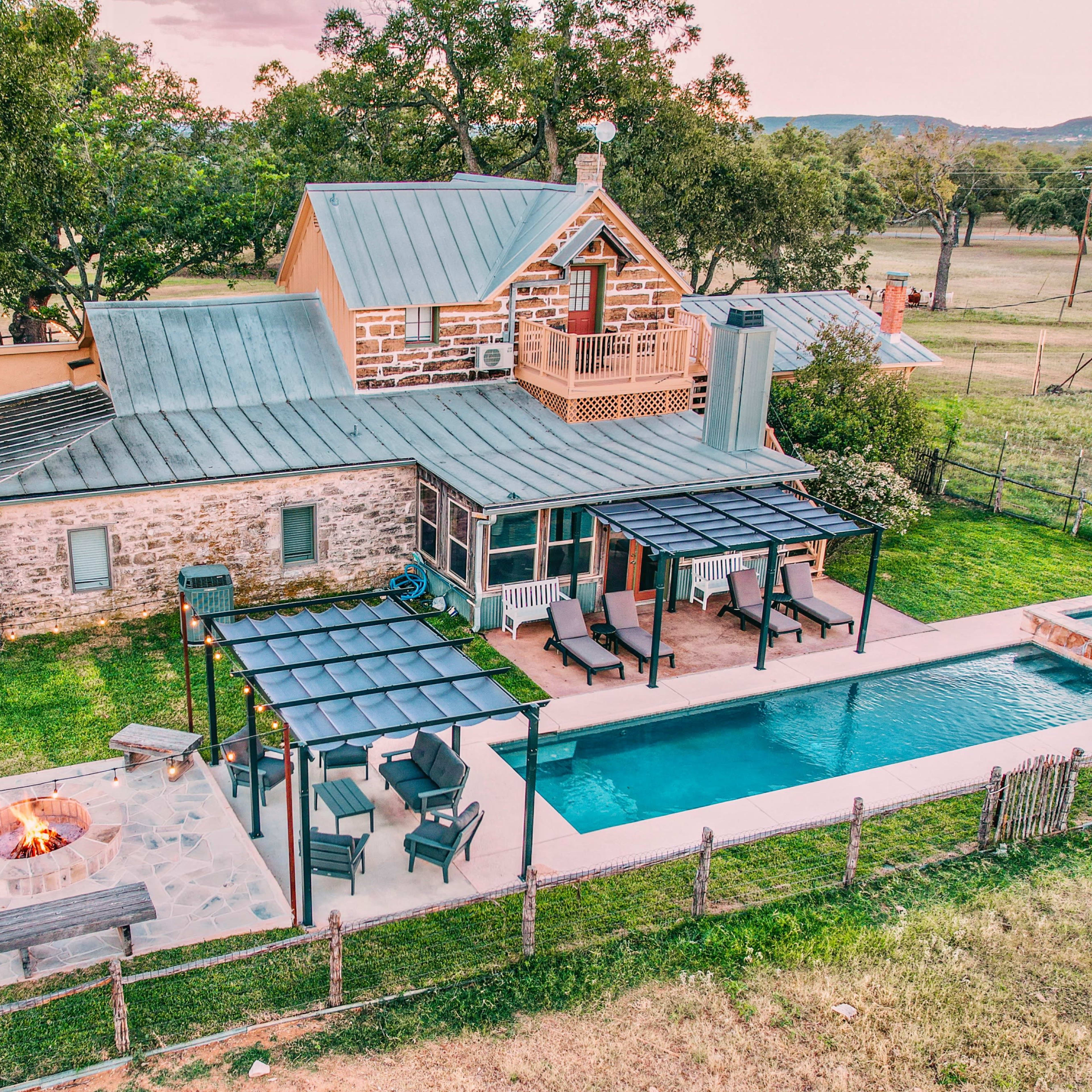 A modern house with a stone exterior, a swimming pool, a fire pit area, and a fenced backyard surrounded by trees.