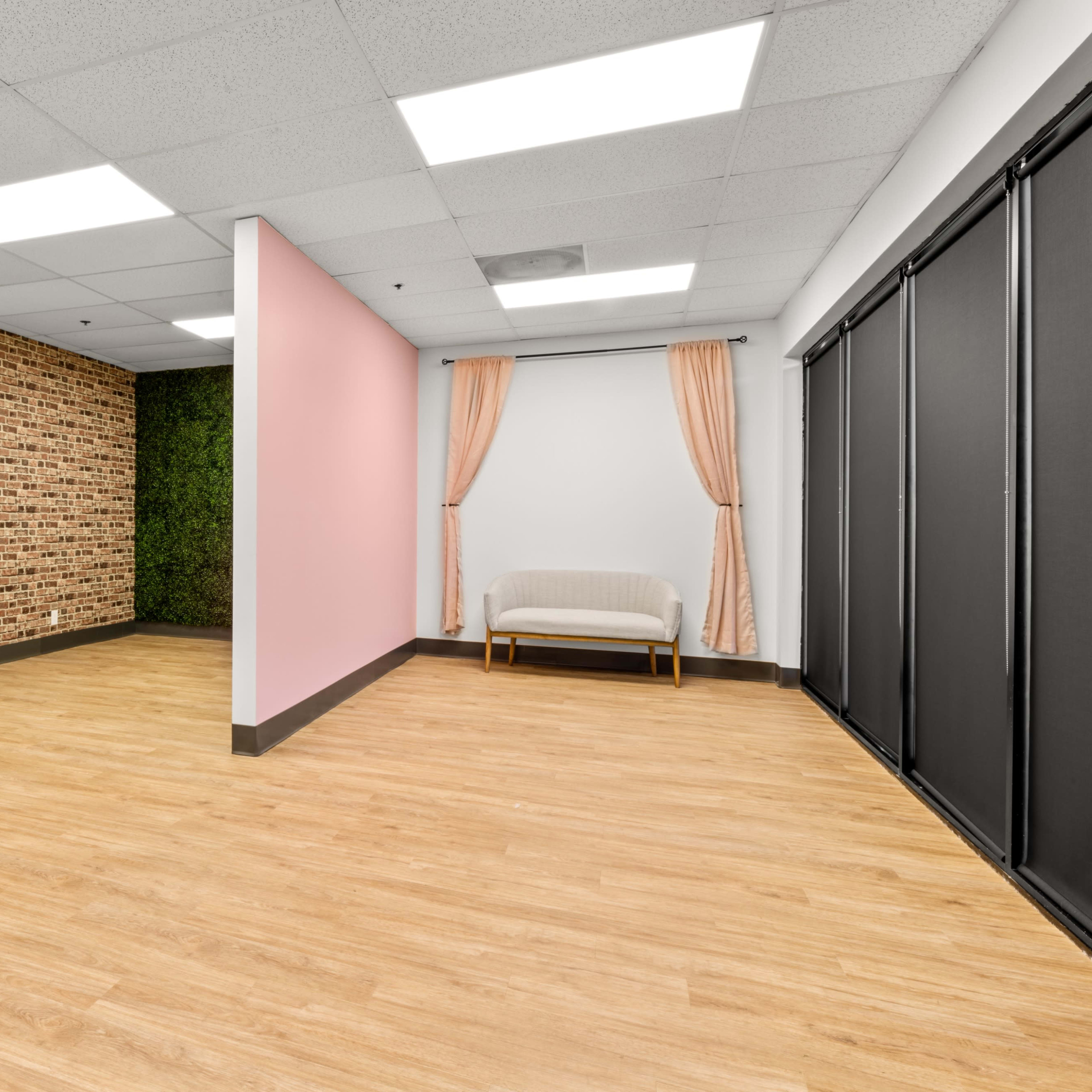 A spacious interior room with one wall featuring exposed brick, another painted pink, and a green wall of plants, complemented by a seating area.