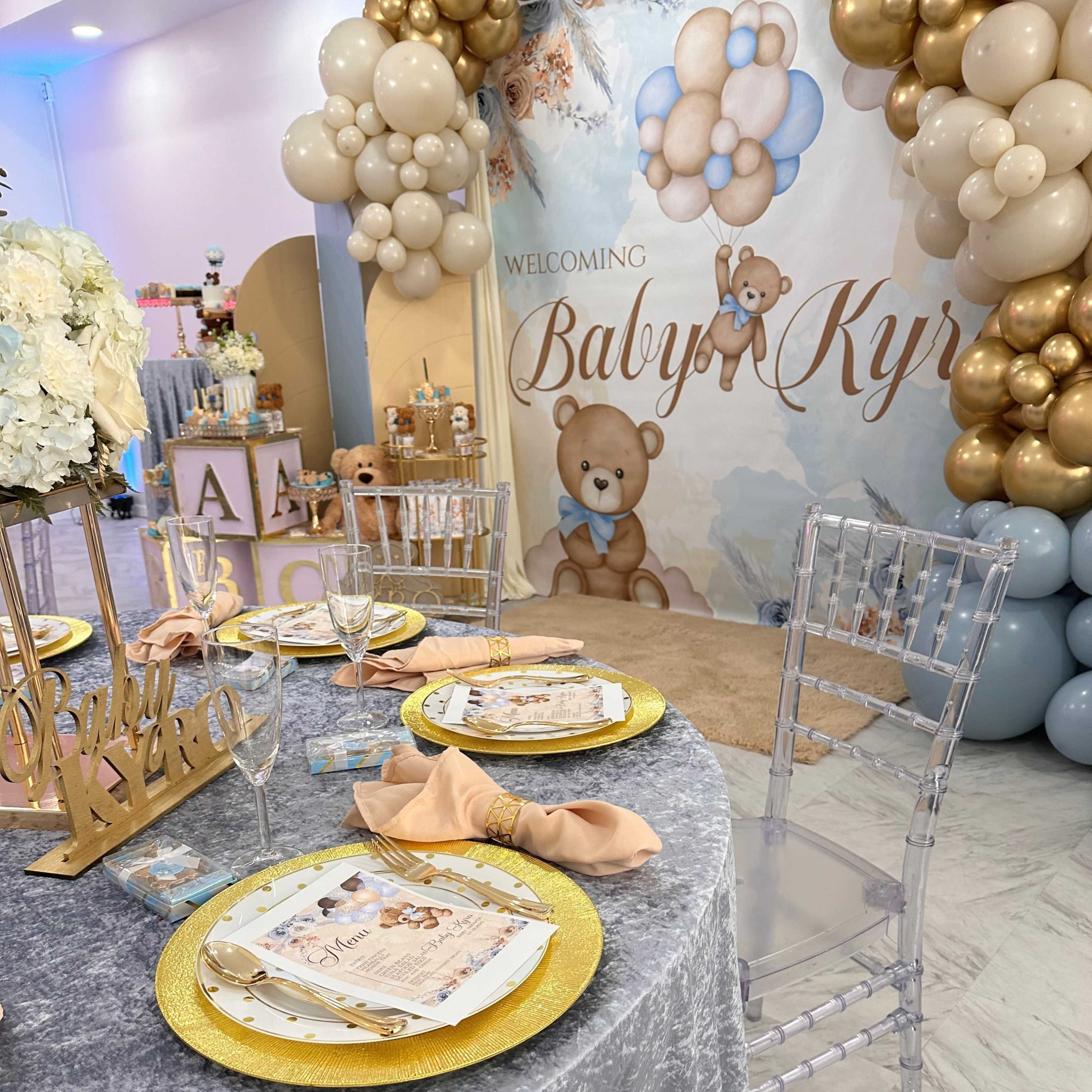 A decorated table for a baby shower, featuring a gray tablecloth, gold and clear place settings, and a backdrop with balloons and a teddy bear theme.