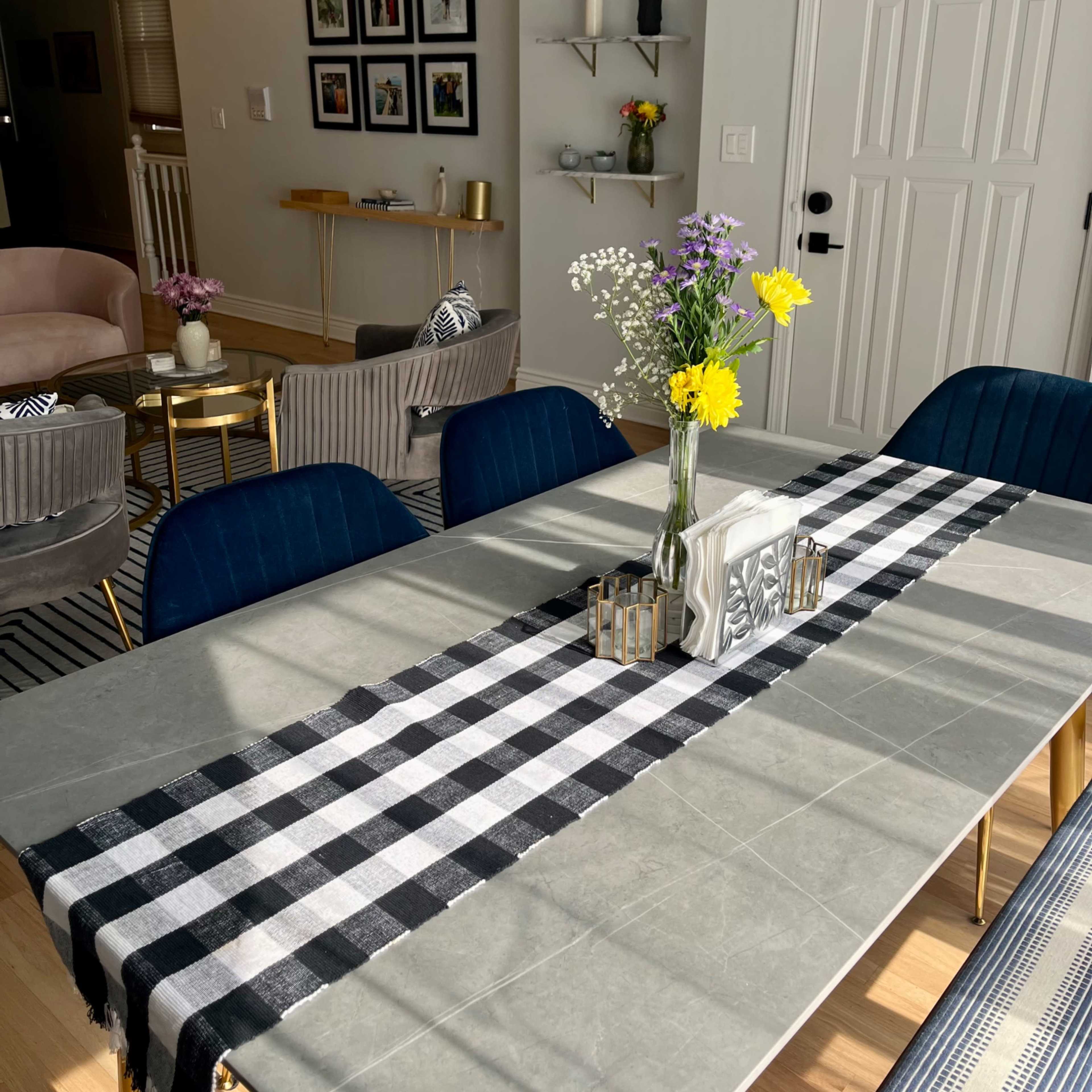 A dining table set with a black and white checked runner is adorned with a vase of flowers, against a backdrop of framed photos on the wall and a cozy living area.