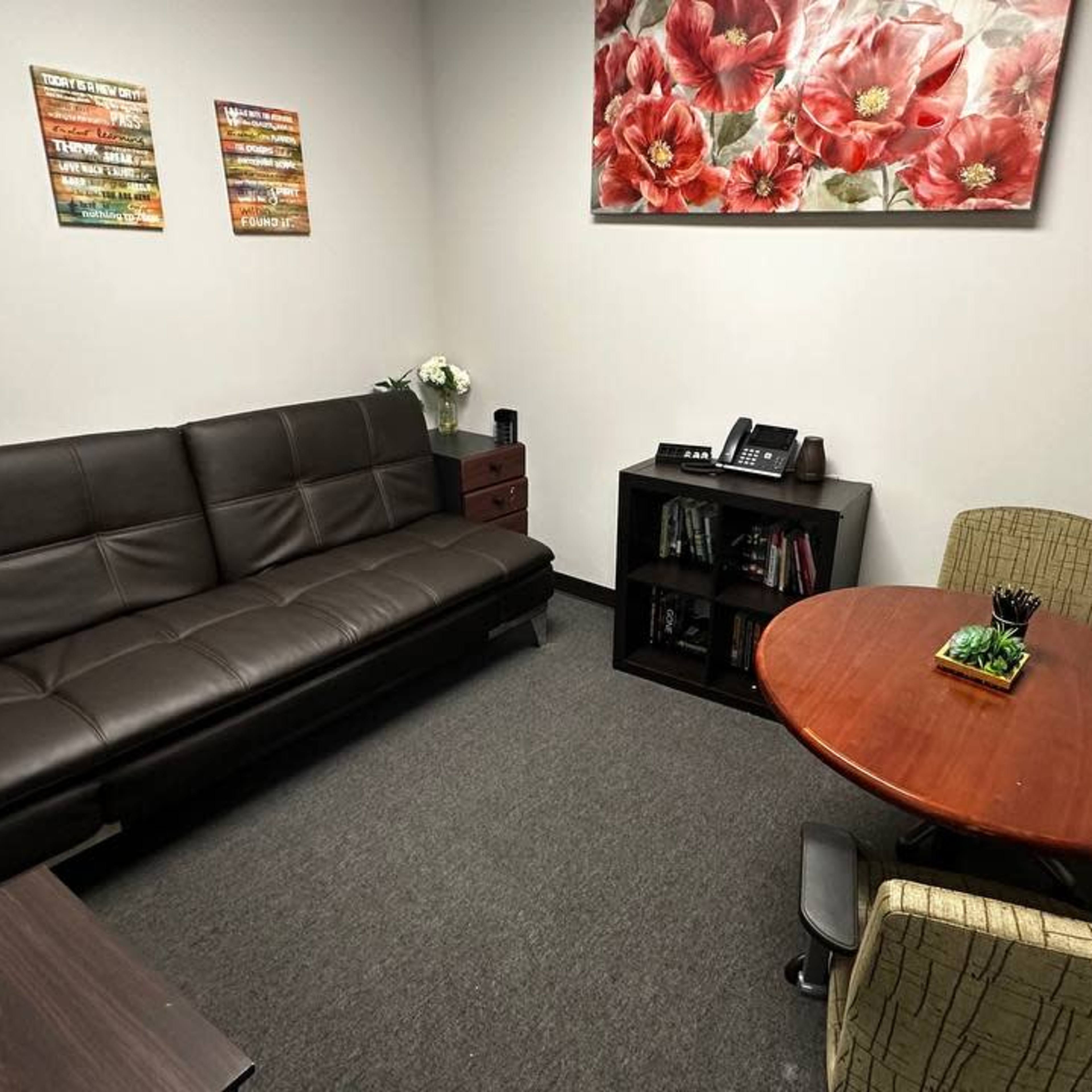 A small, neatly arranged office with a brown sofa, a round table with a plant, and a wall adorned with floral artwork.