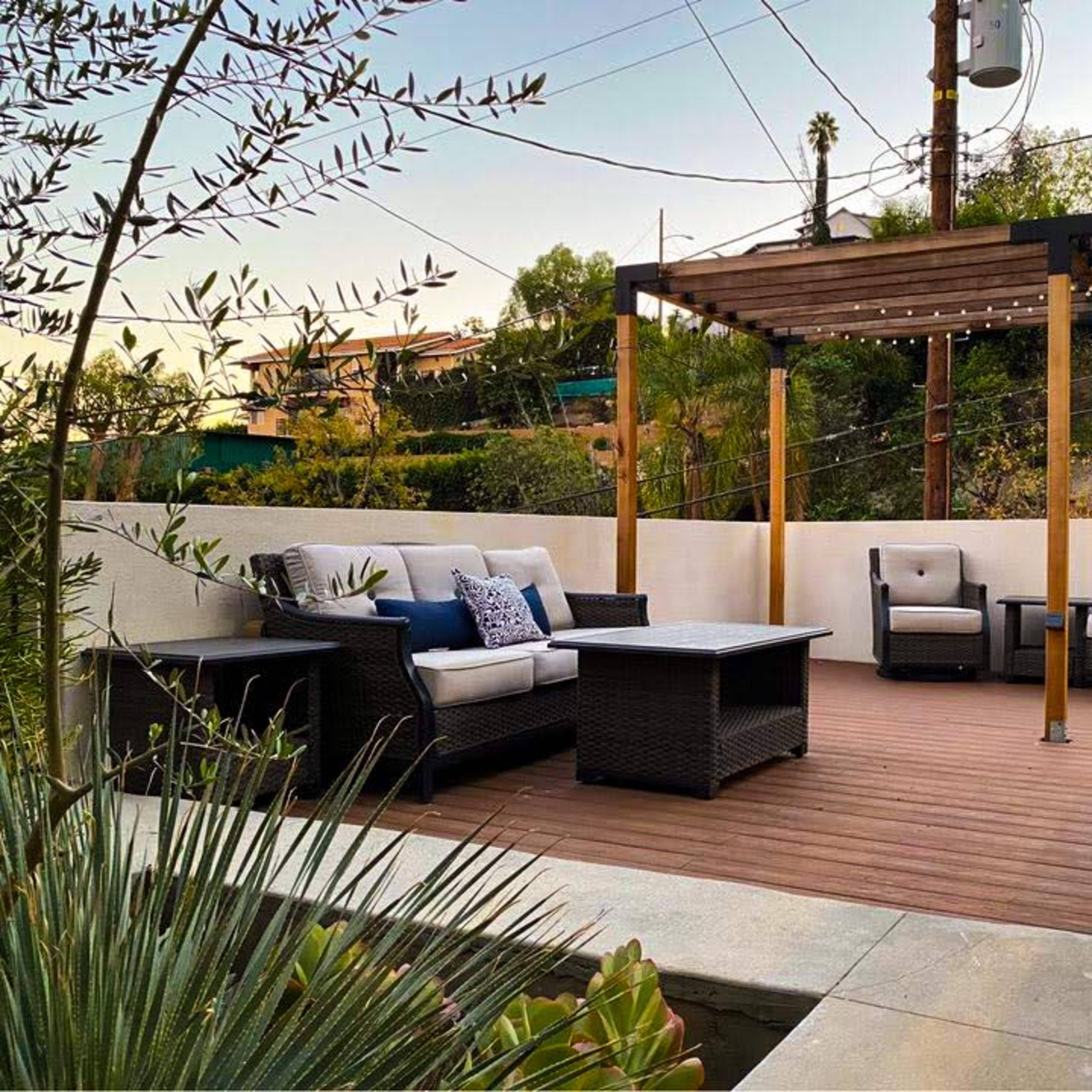 A wooden deck with outdoor seating and a pergola is positioned in a landscaped area surrounded by greenery.