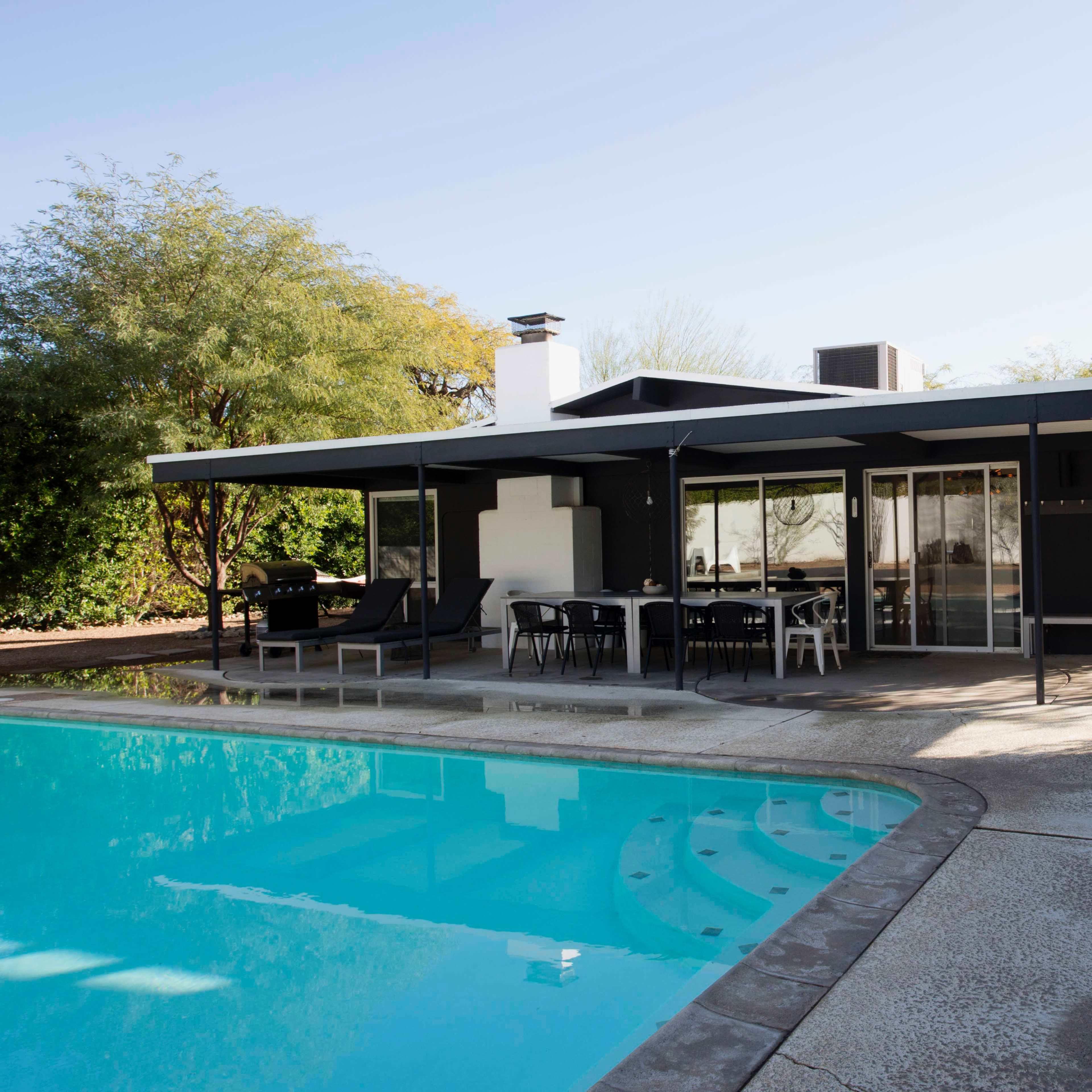 A modern house with a flat roof and large glass windows overlooks a swimming pool surrounded by a concrete patio.