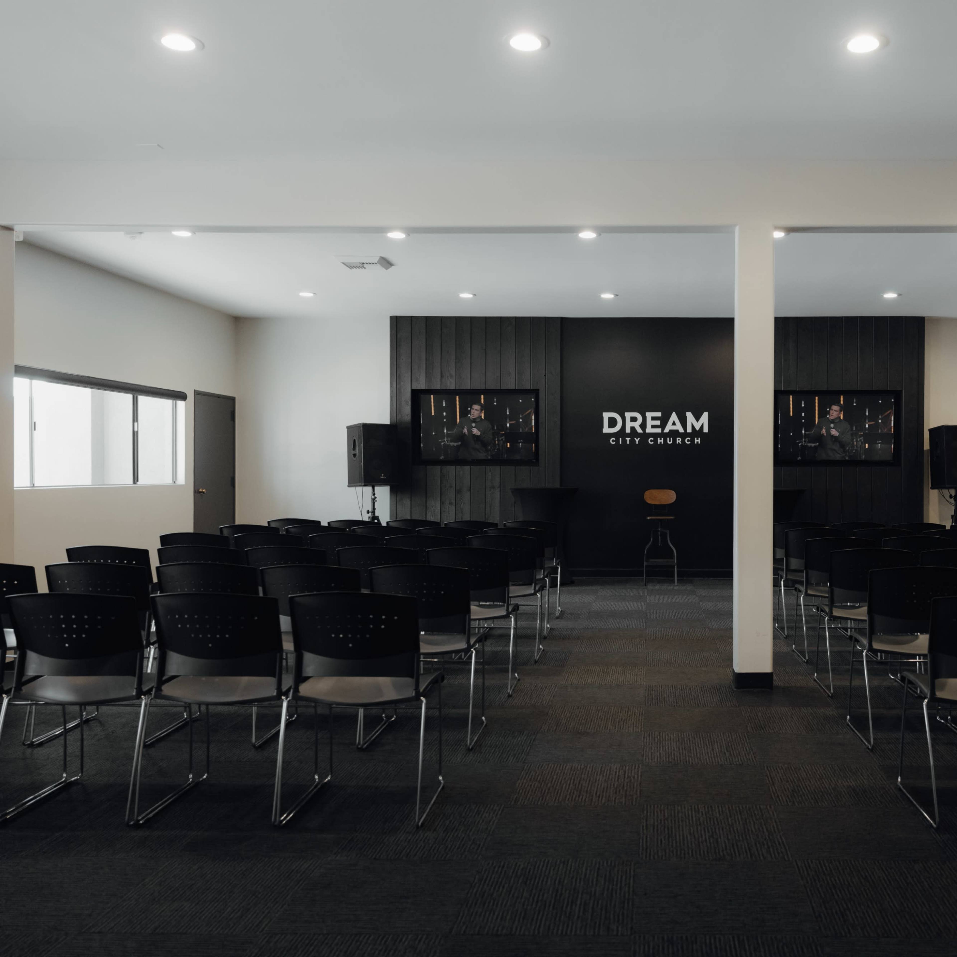 A conference room features rows of black chairs facing a dark wall with the words "DREAM CITY CHURCH" illuminated, flanked by two screens.