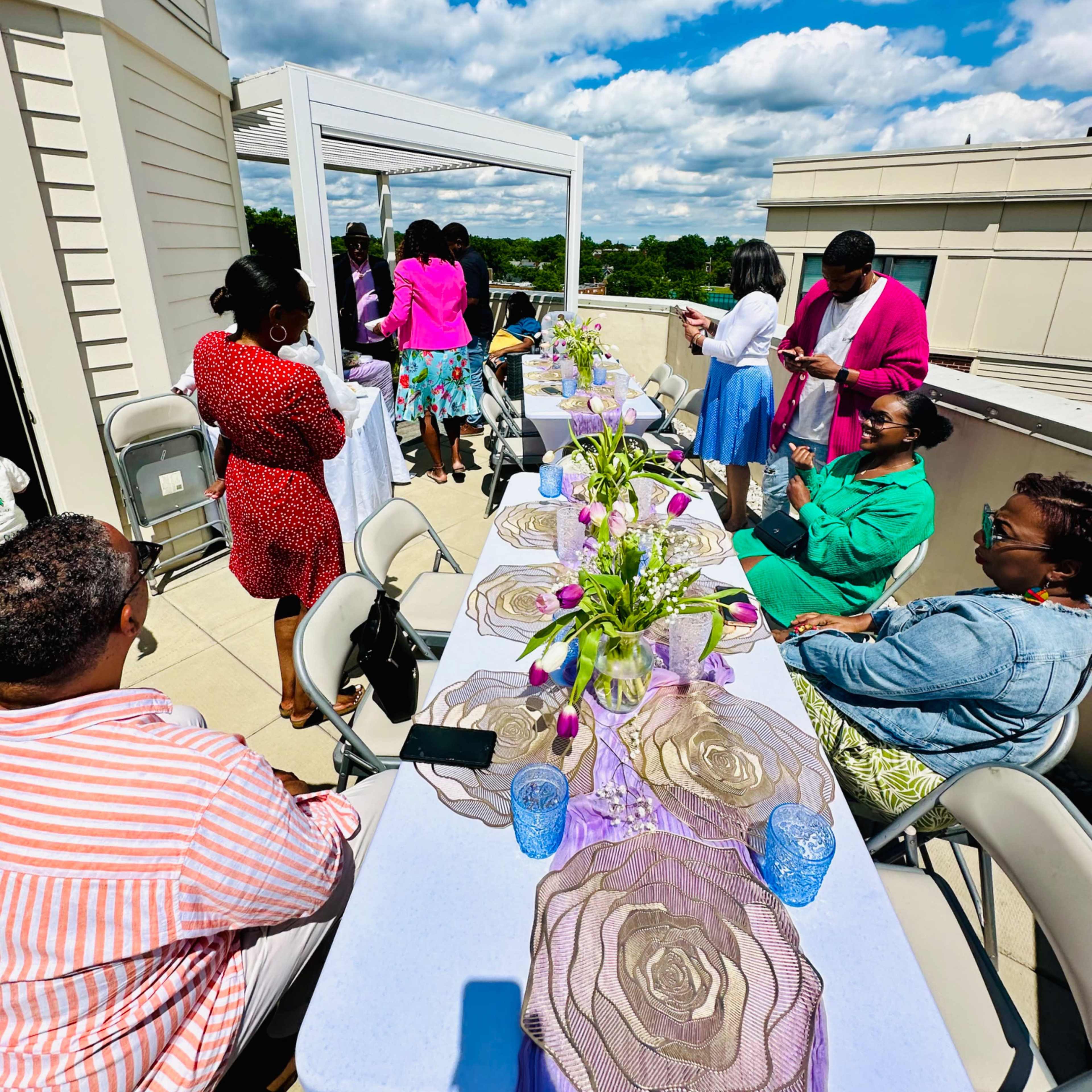 A group of people gather around decorated tables on a rooftop, enjoying a sunny day with flowers and glassware.