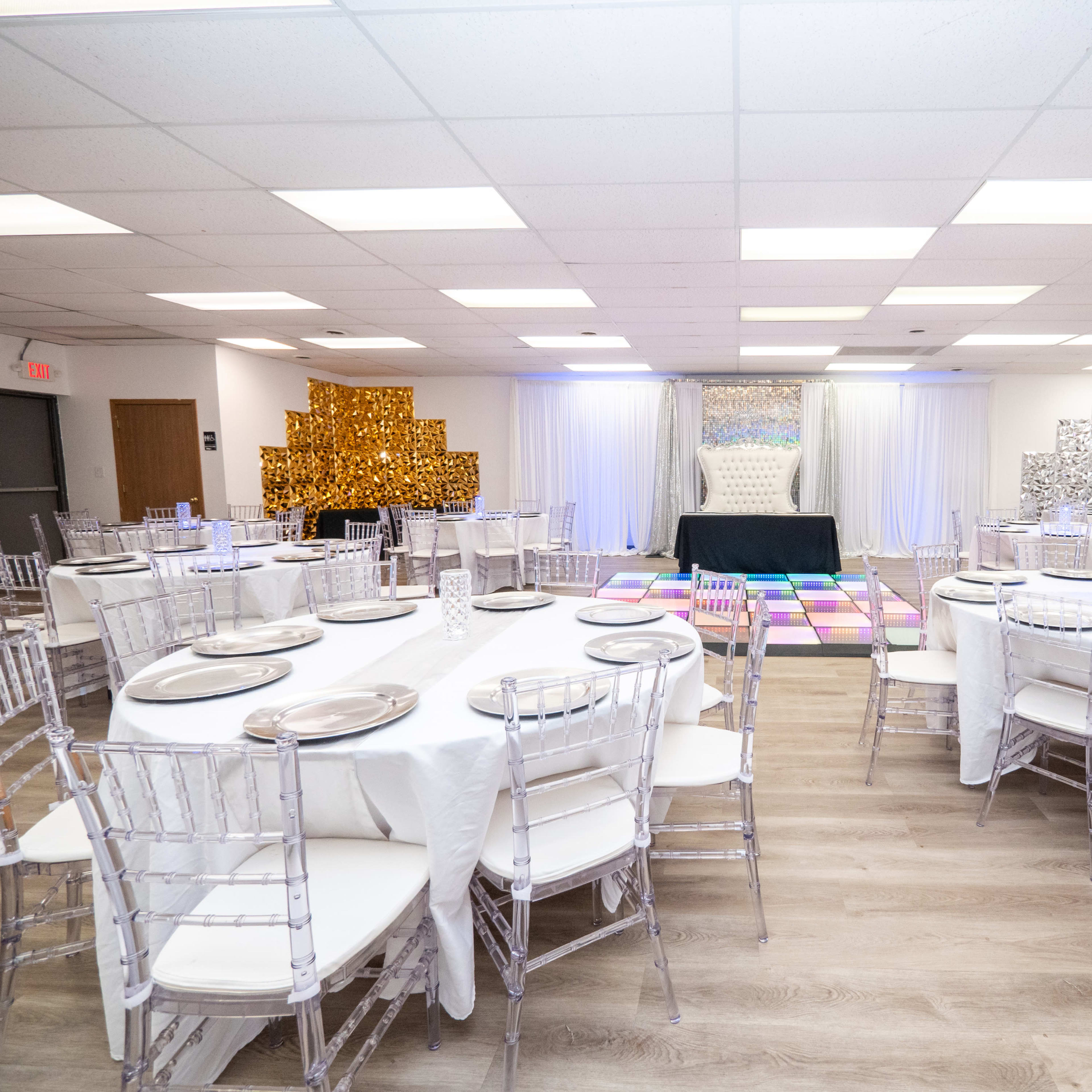 A banquet hall set up with round tables and clear chiavari chairs, featuring decorative backdrops on the walls and a central stage area.