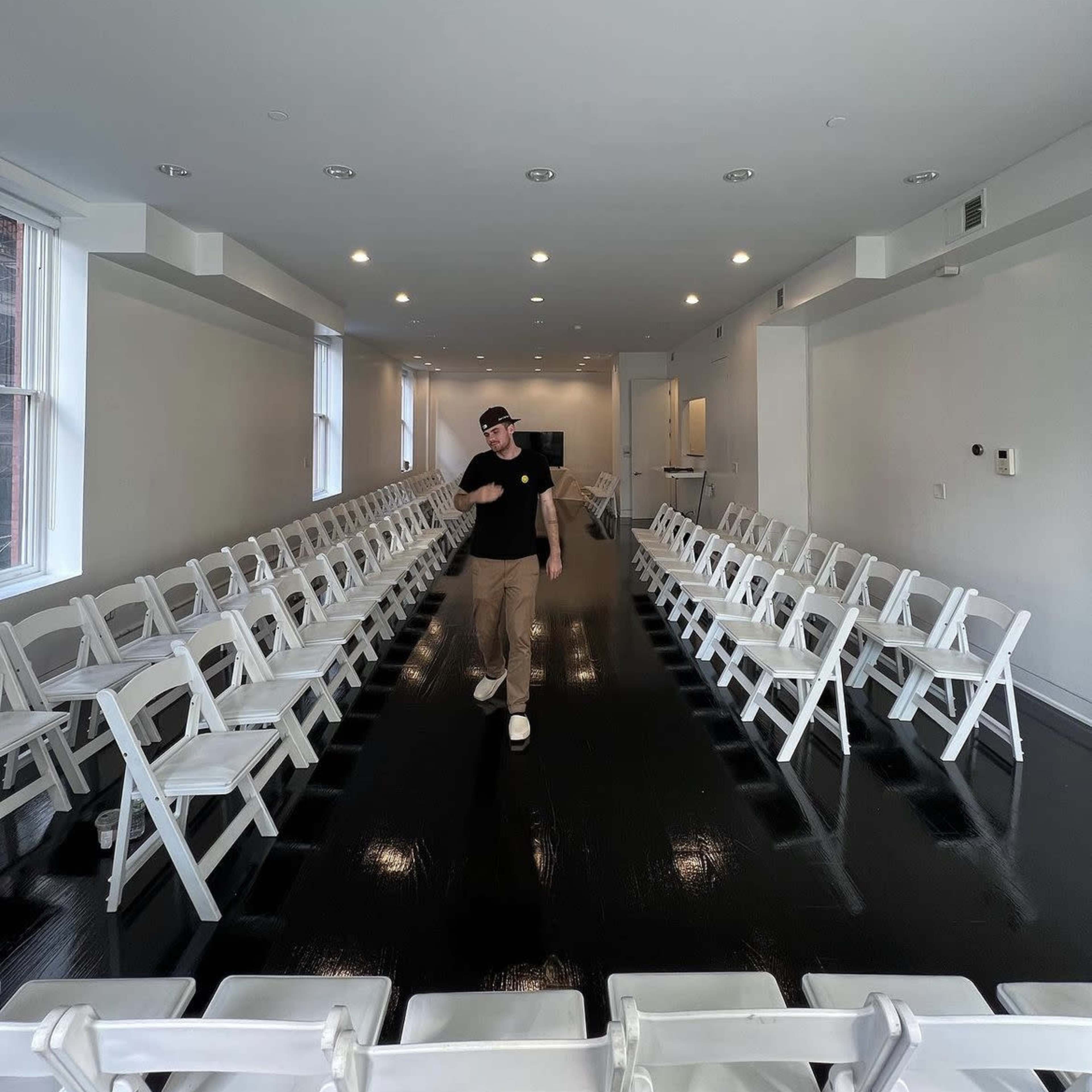 A man walks through a room arranged with rows of white chairs on a polished black floor.