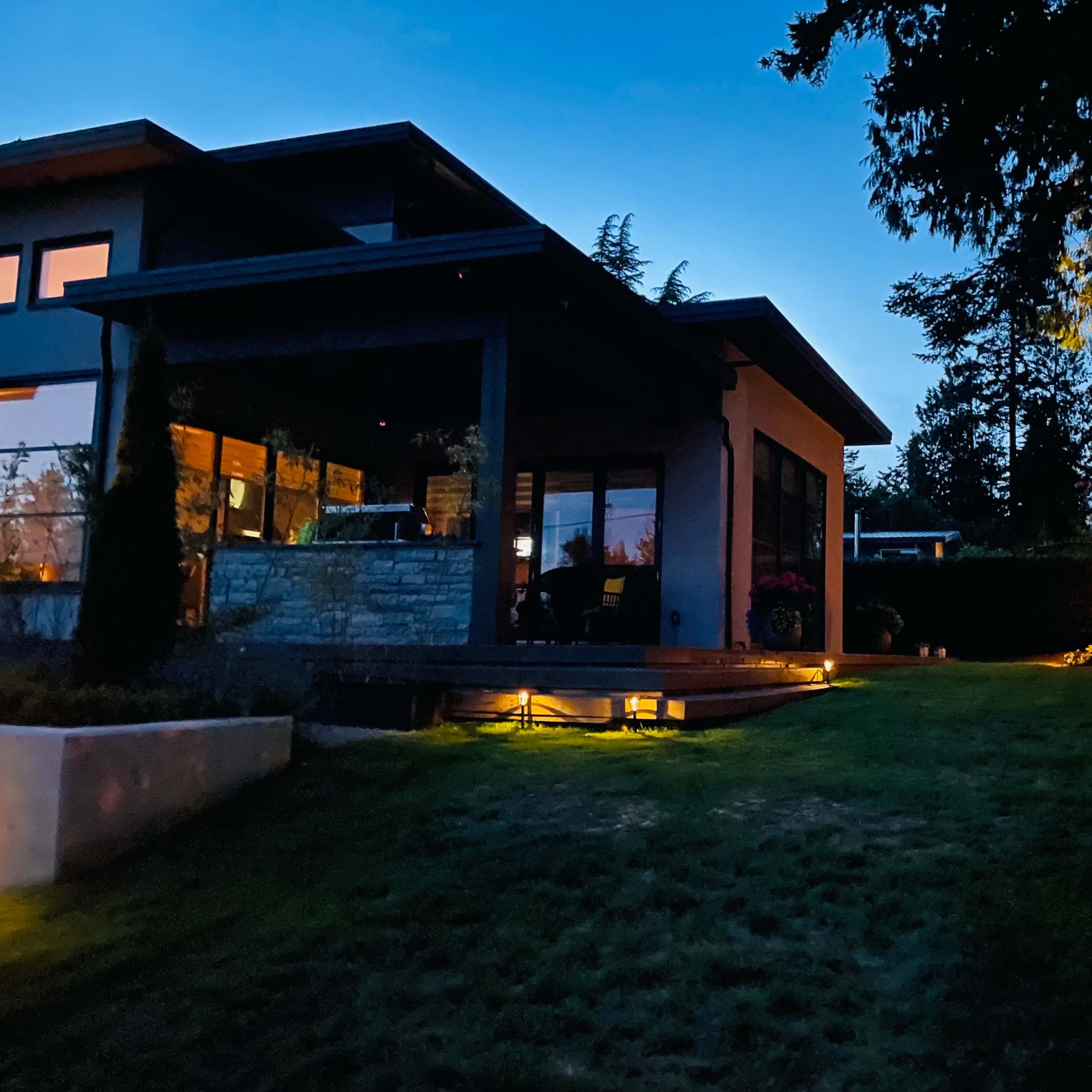 A modern house with outdoor lighting illuminating the garden and patio area during dusk.