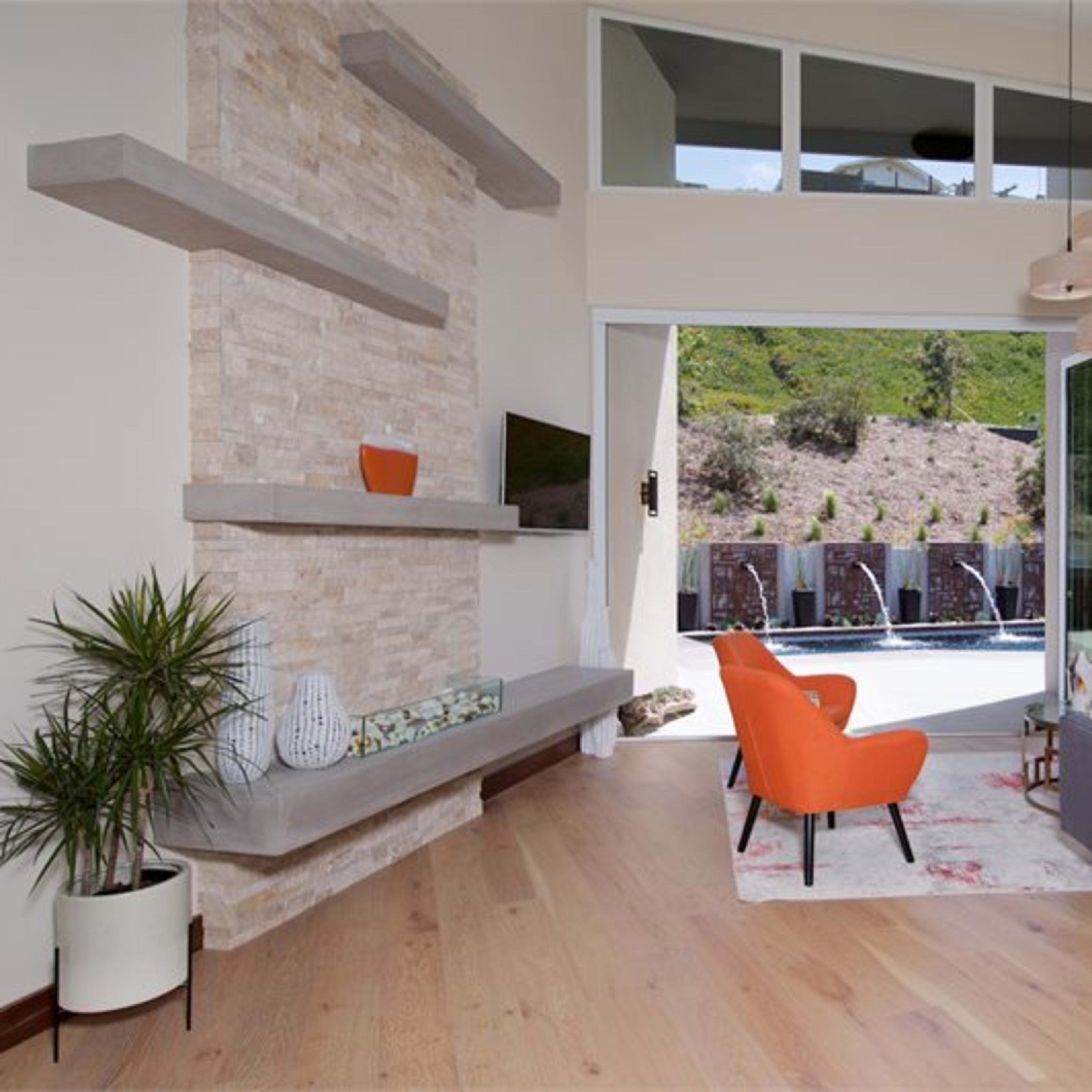 A modern living room features a stone fireplace, large windows, and a patio overlooking a pool.
