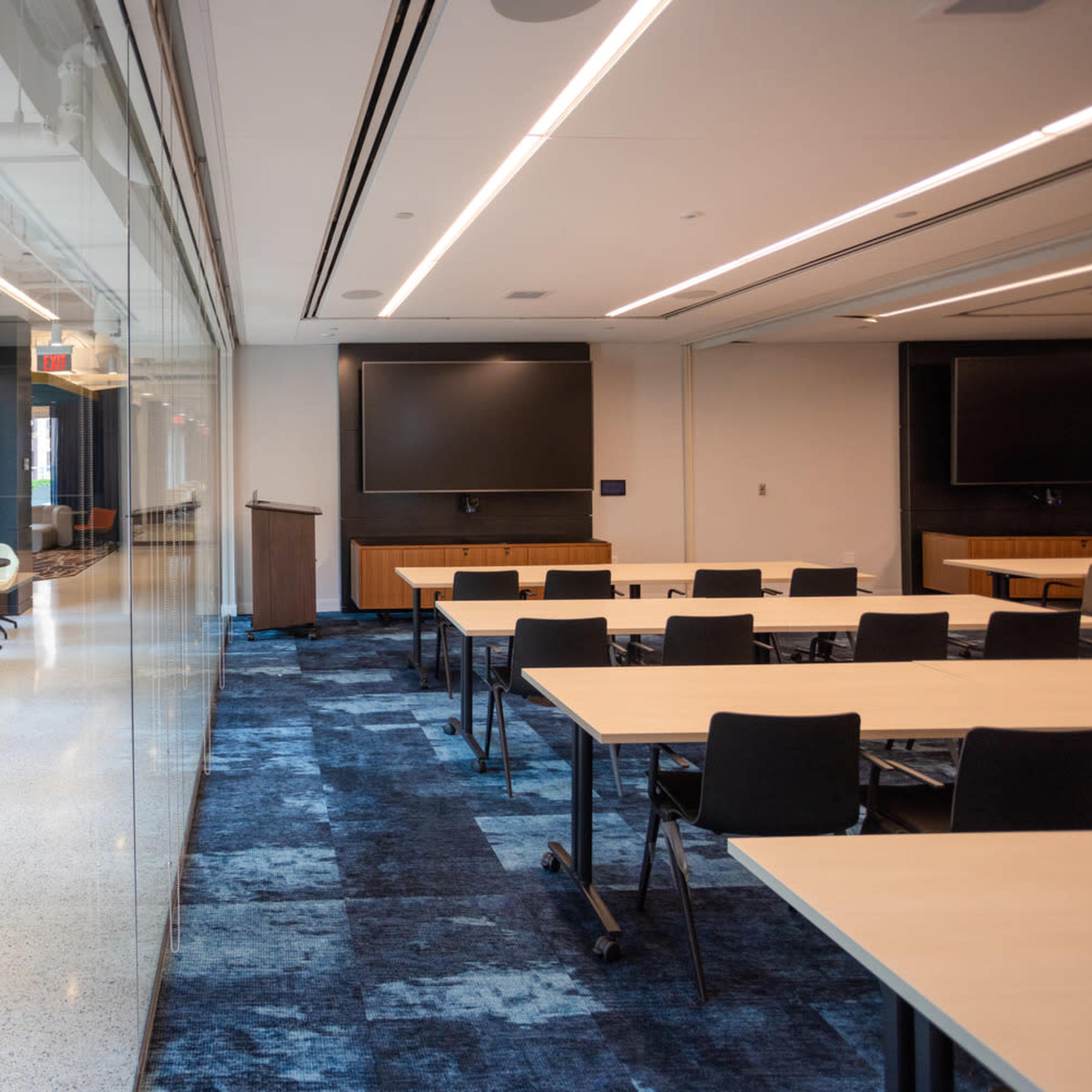 A modern conference room with glass walls, rows of tables and chairs, and large screens on the opposite wall.