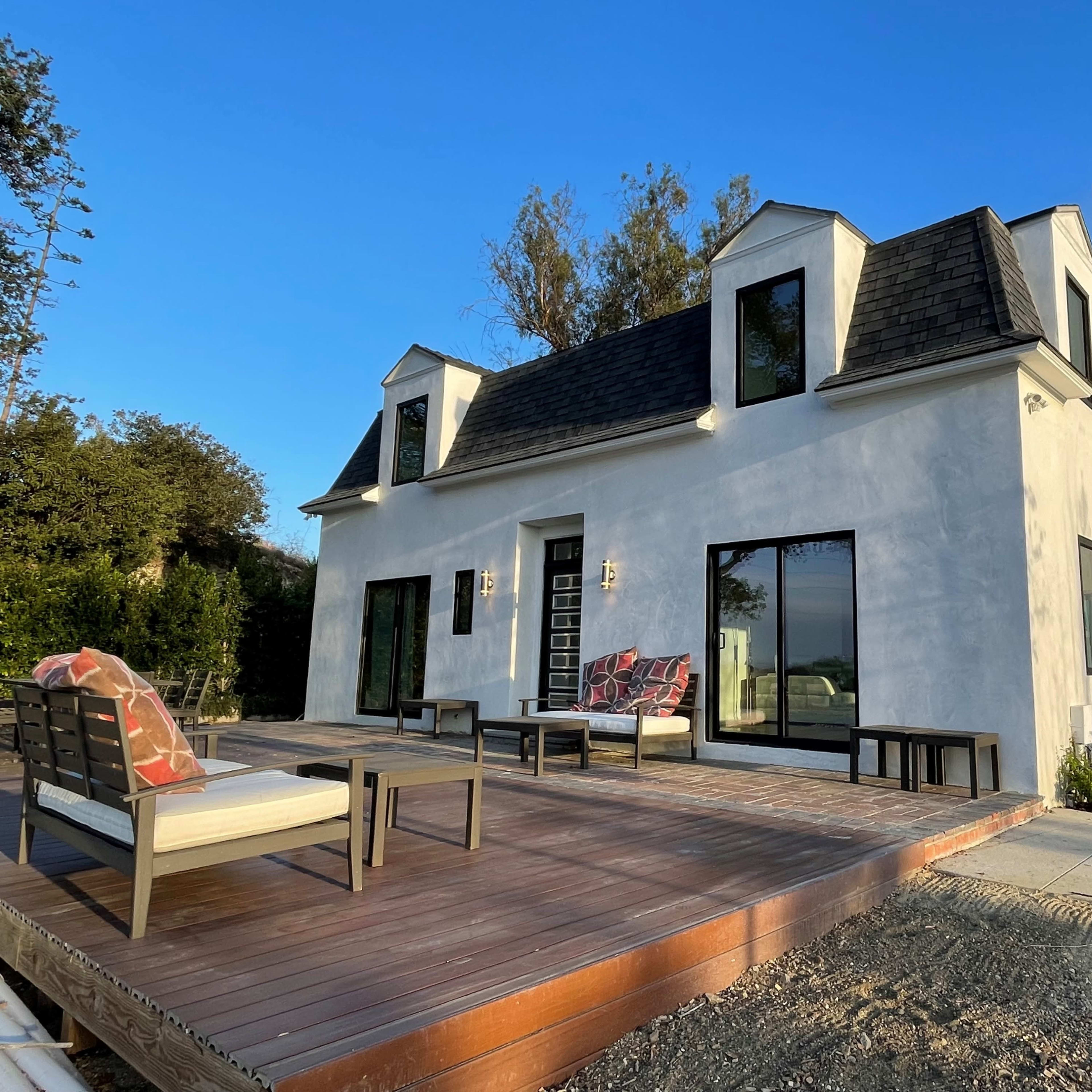 A modern two-story house with a sloped roof and large windows is situated next to a wooden deck and a leafless tree.
