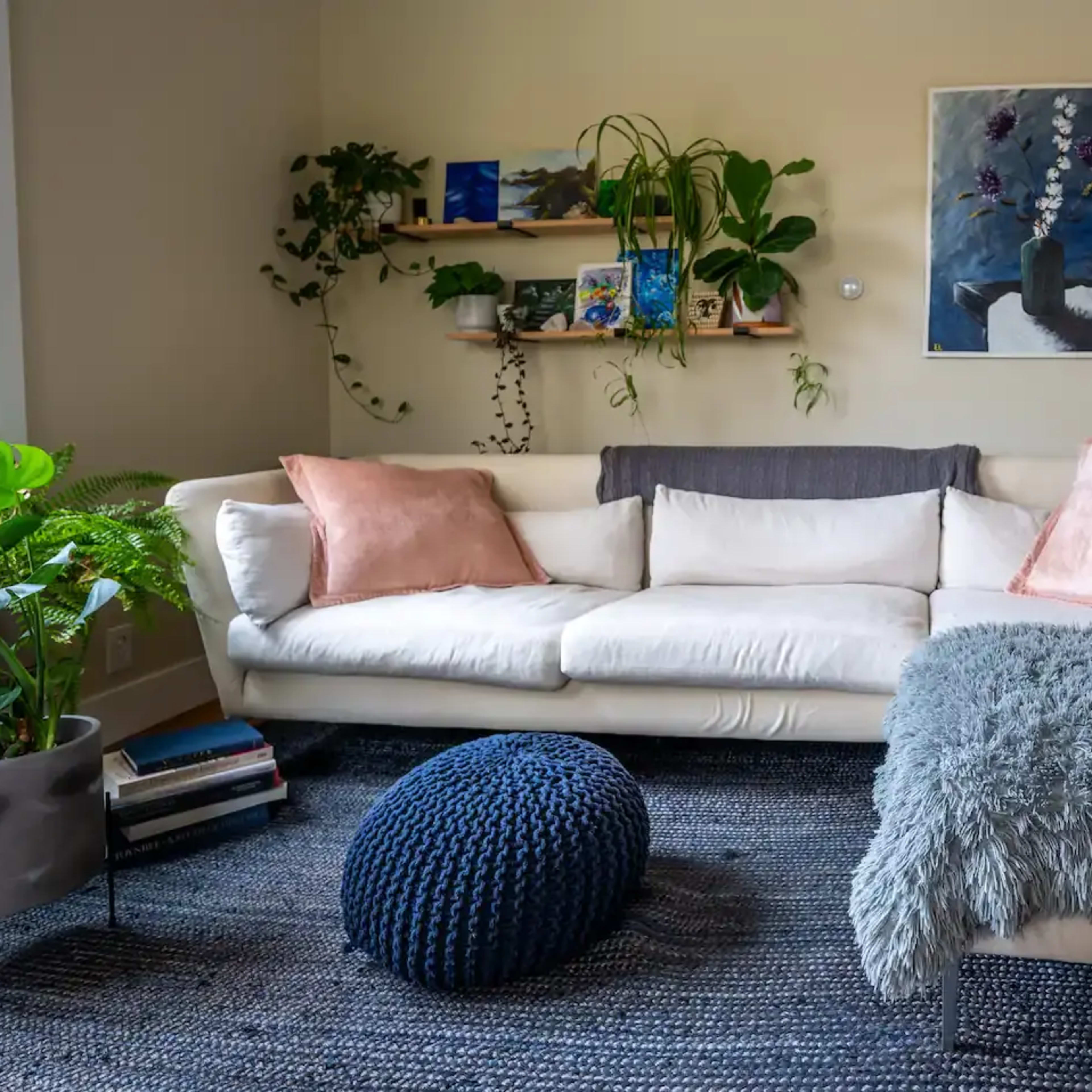 A bright living room features a white sectional sofa with pink pillows, a textured gray throw, and a blue pouf, surrounded by plants and artwork on the walls.