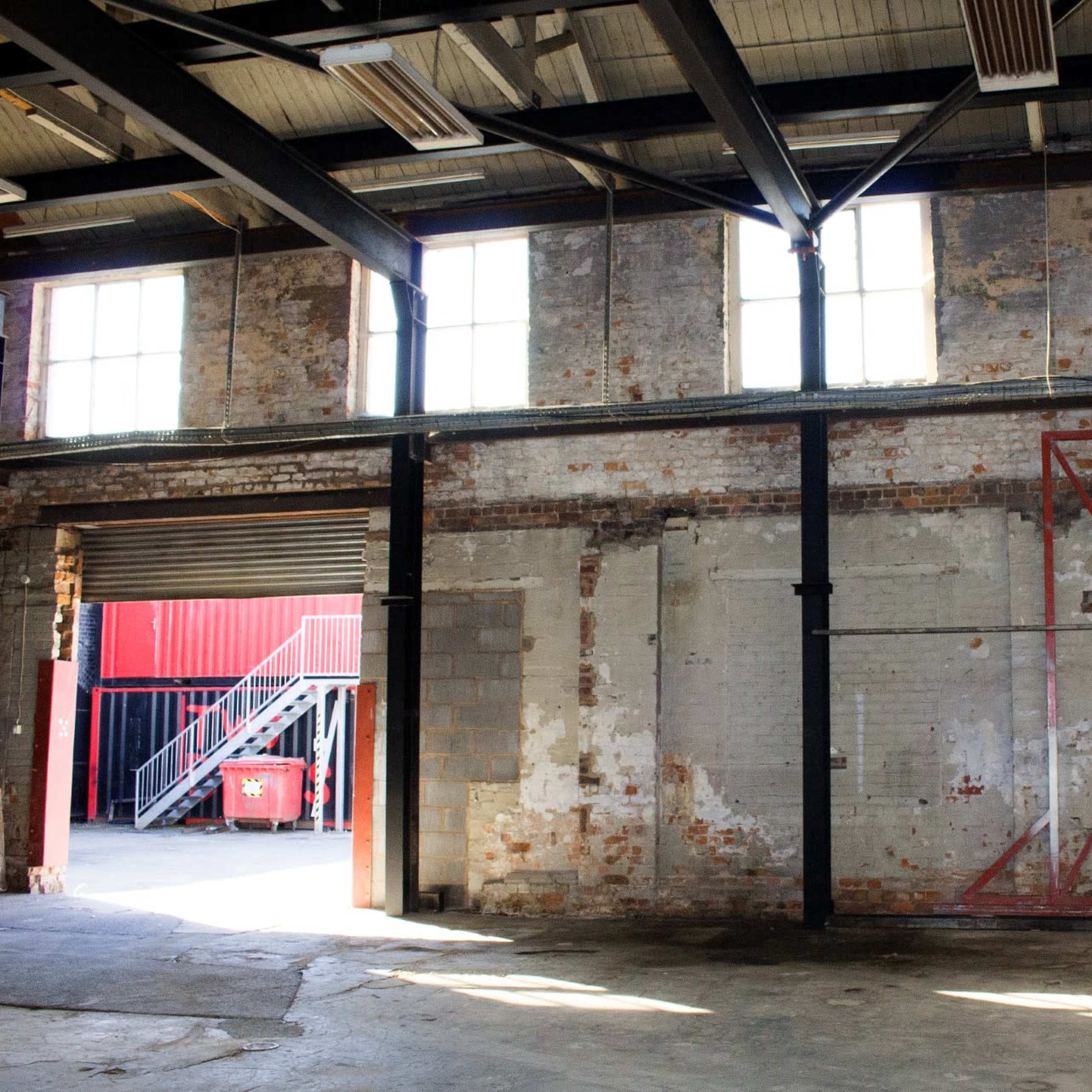 An industrial warehouse interior with exposed brick walls, large windows, and a red door frame.