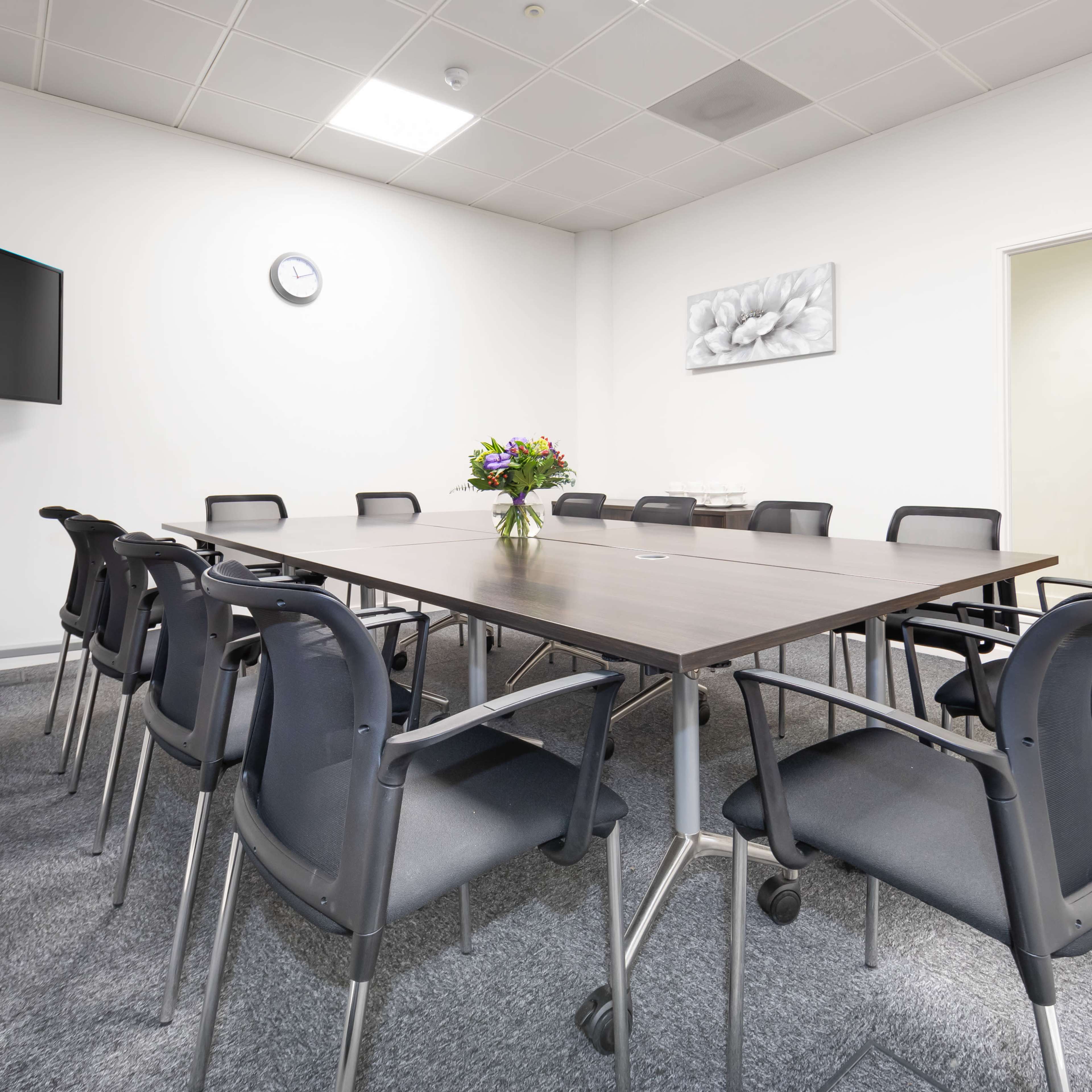 A large, rectangular table surrounded by ten black chairs is positioned in a well-lit conference room with a TV on a stand and a floral arrangement in the center.