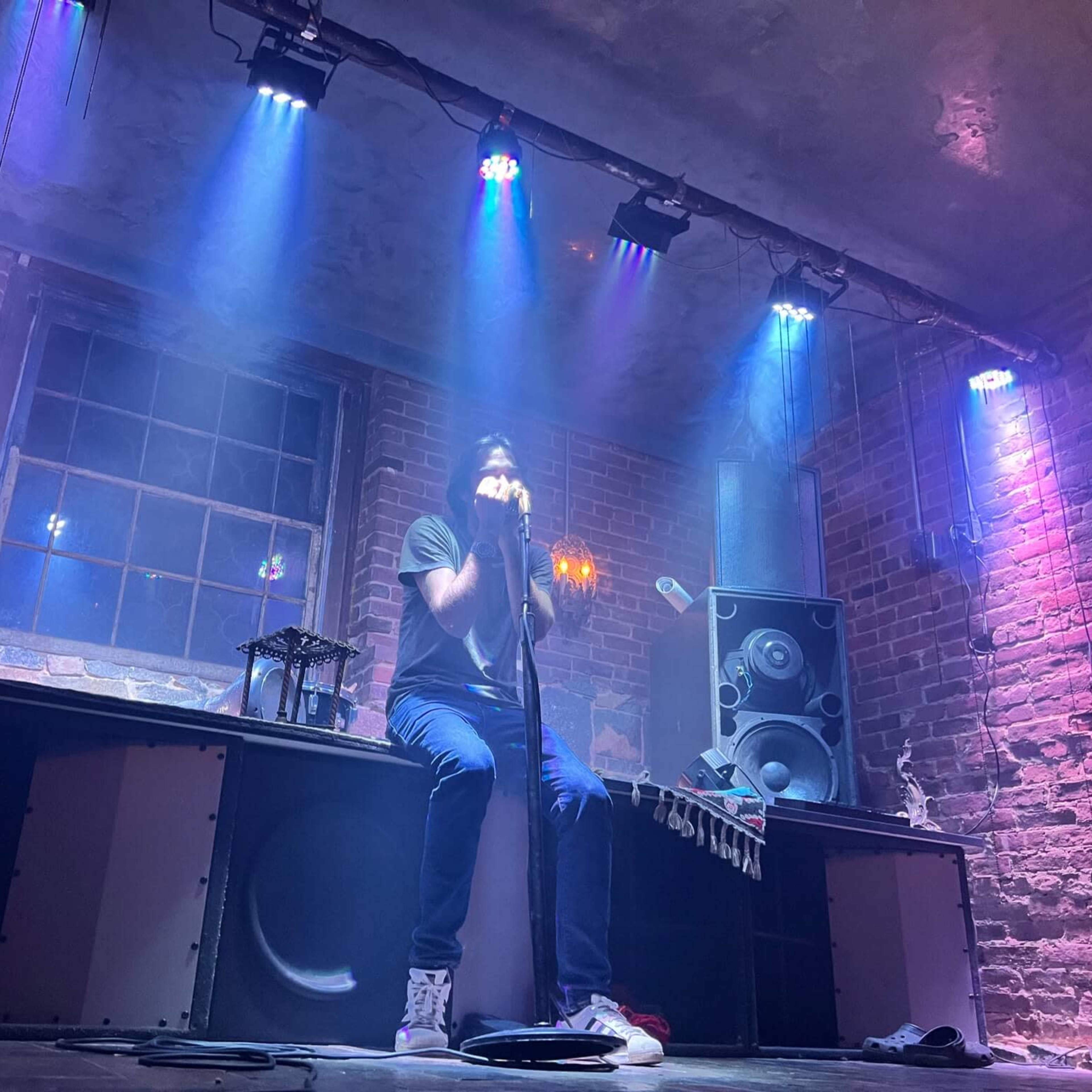 A person is singing into a microphone on a stage surrounded by colorful lights and a sound system in a brick-walled room.