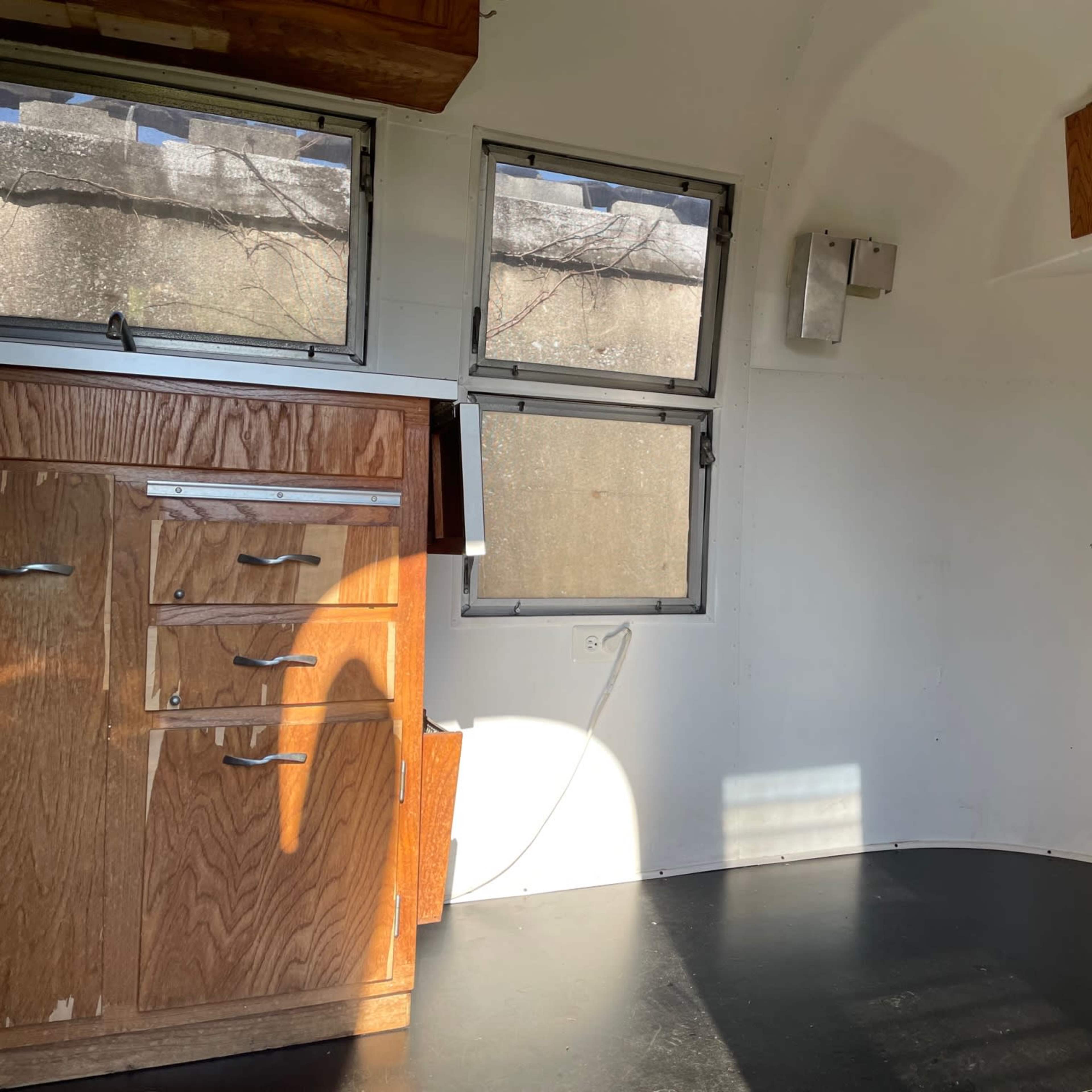 The interior of a small trailer features wooden cabinetry, a counter space, and large windows with natural light entering the room.
