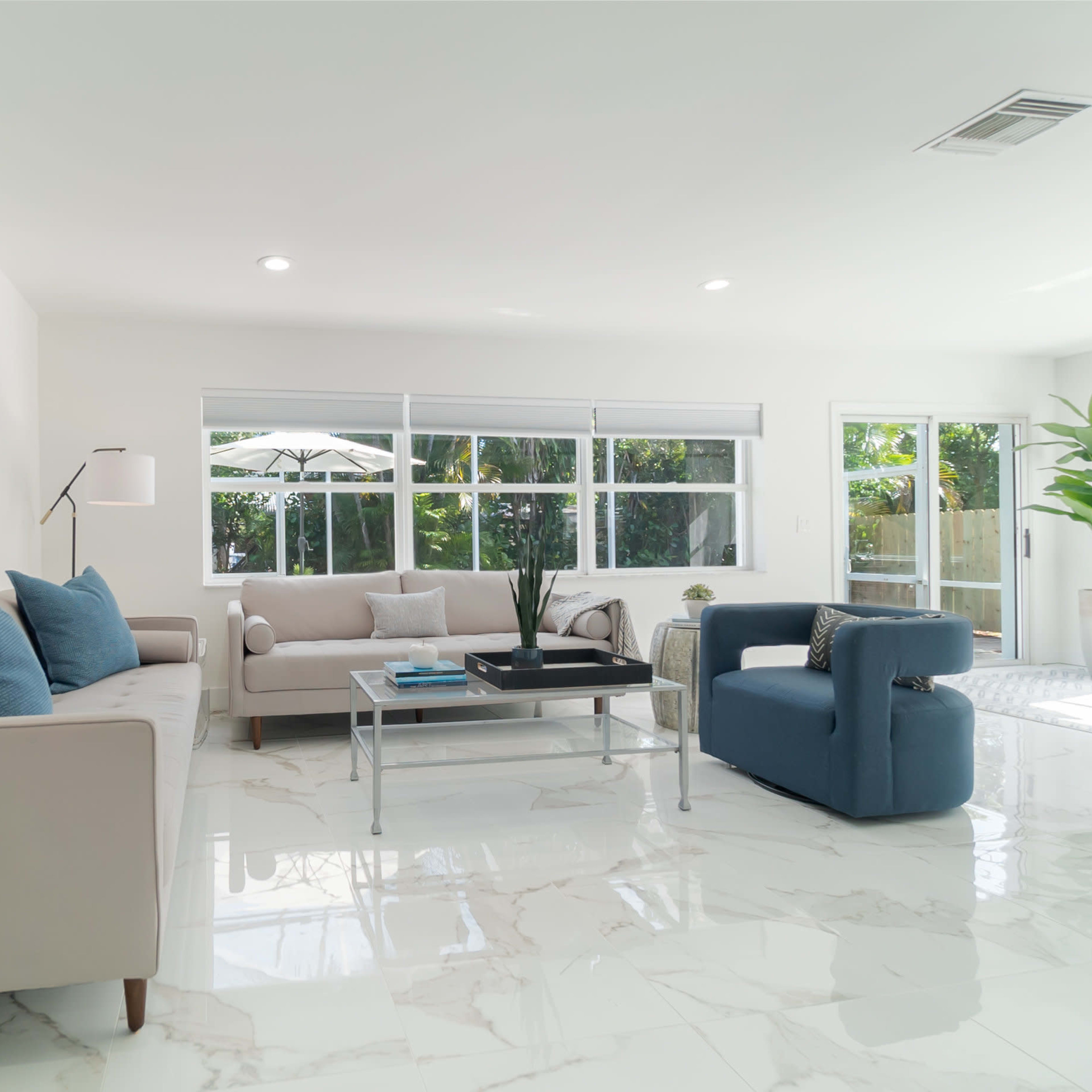 A modern living room features a light-colored sofa, a blue armchair, and large windows that overlook a patio with greenery.
