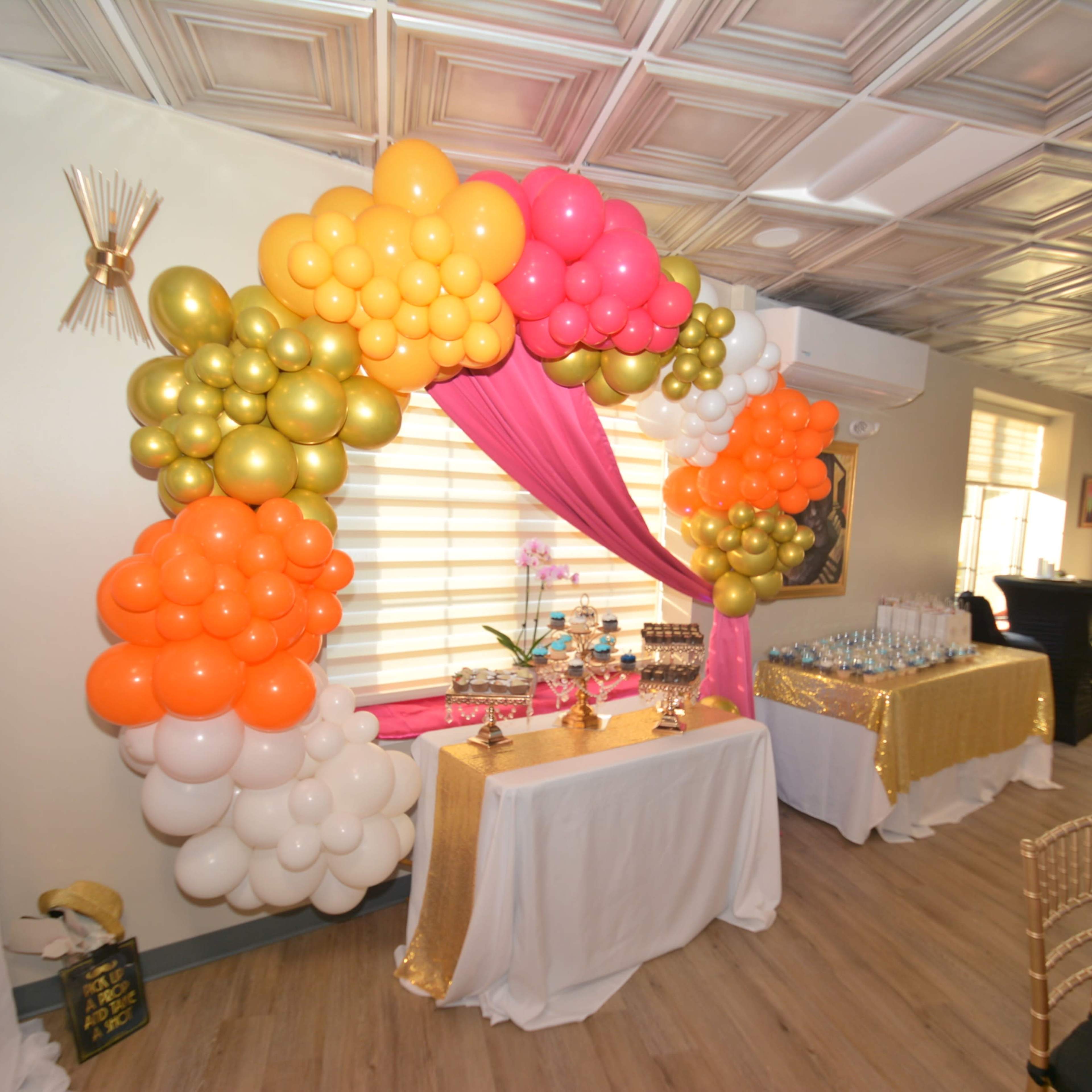 A balloon arch with orange, pink, and gold balloons is set above a decorated table with snacks and drinks in a party room.