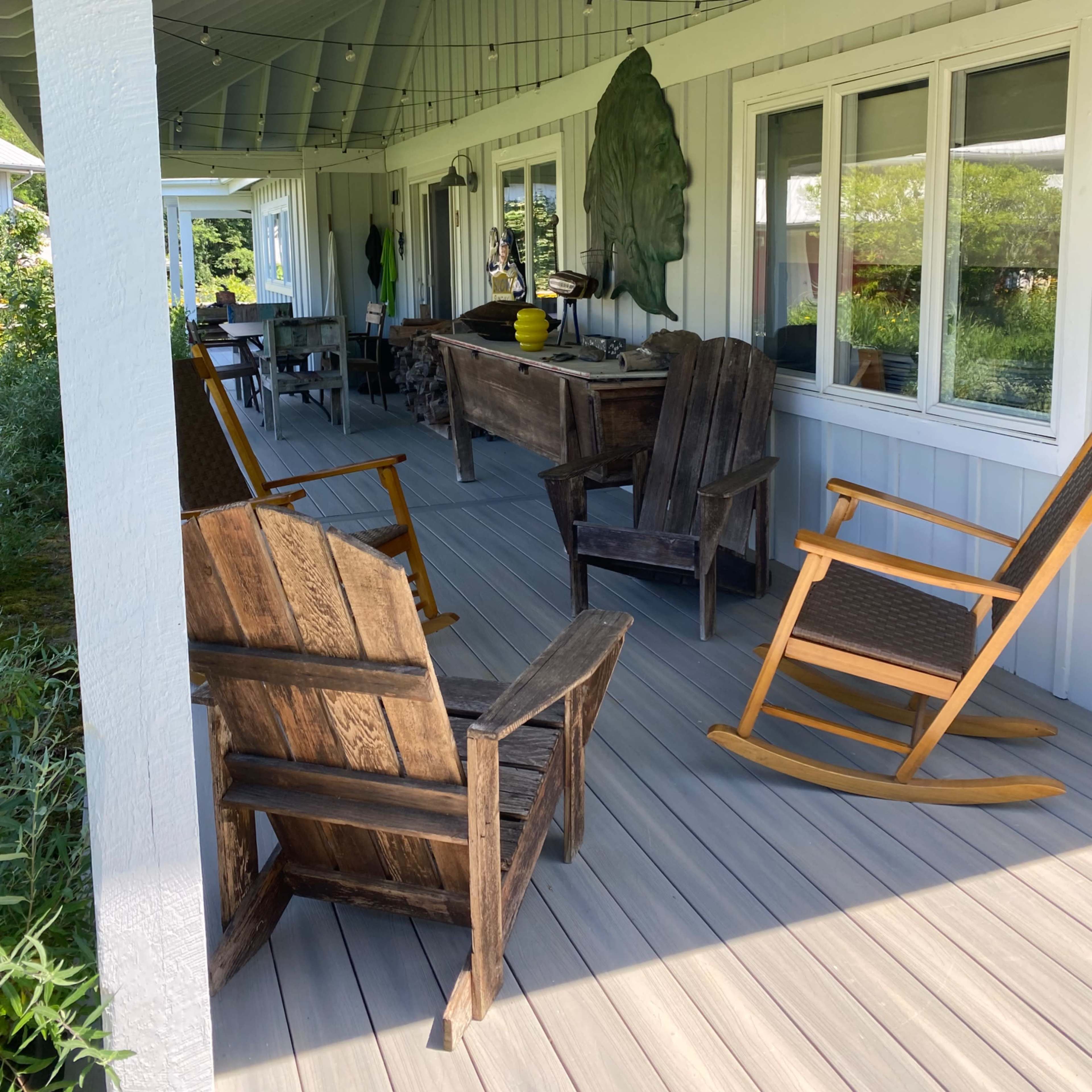 A covered porch features four wooden chairs and a wooden table, with string lights and a view of a garden beyond.