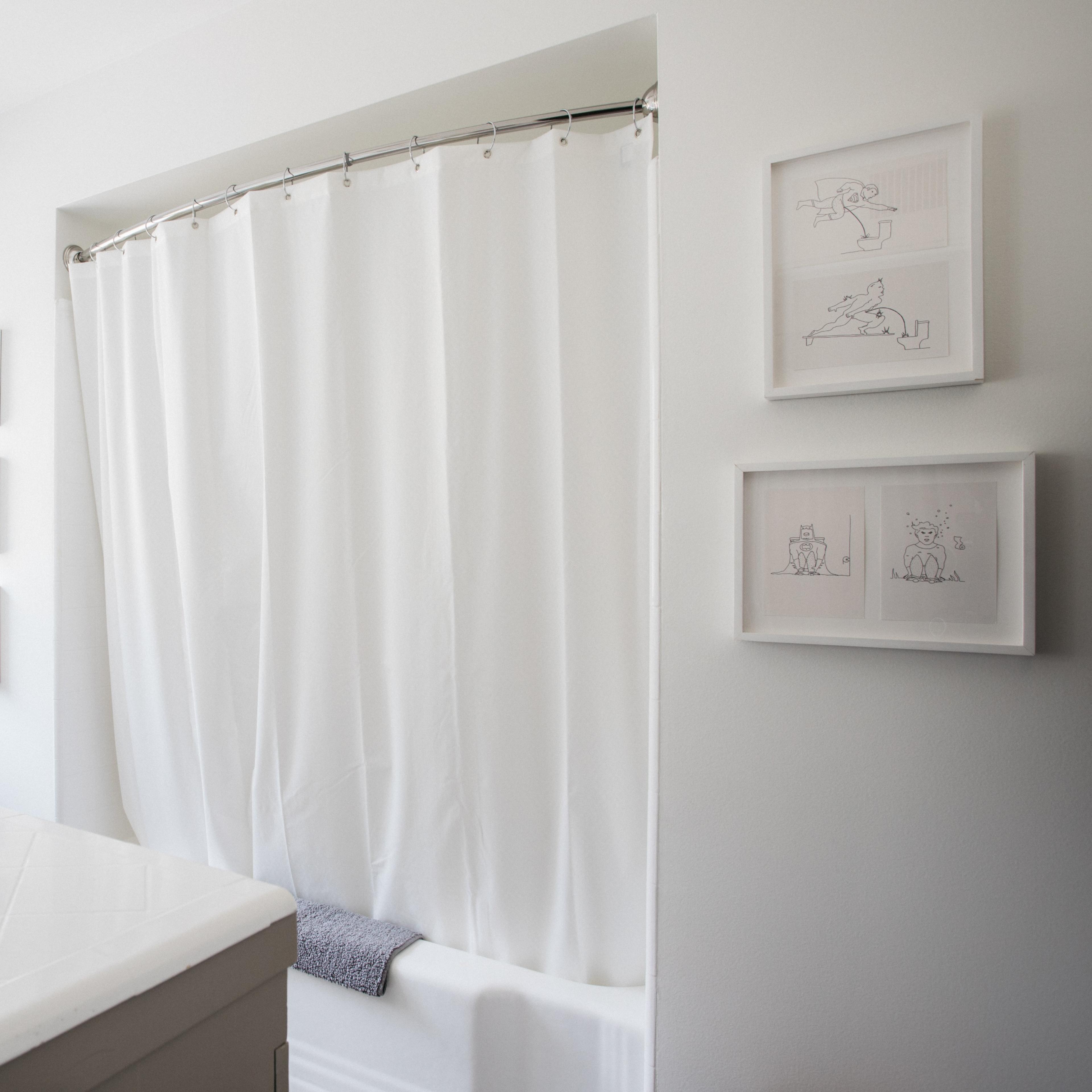 A bright bathroom features a white shower curtain, a window for natural light, and framed artworks on the wall.