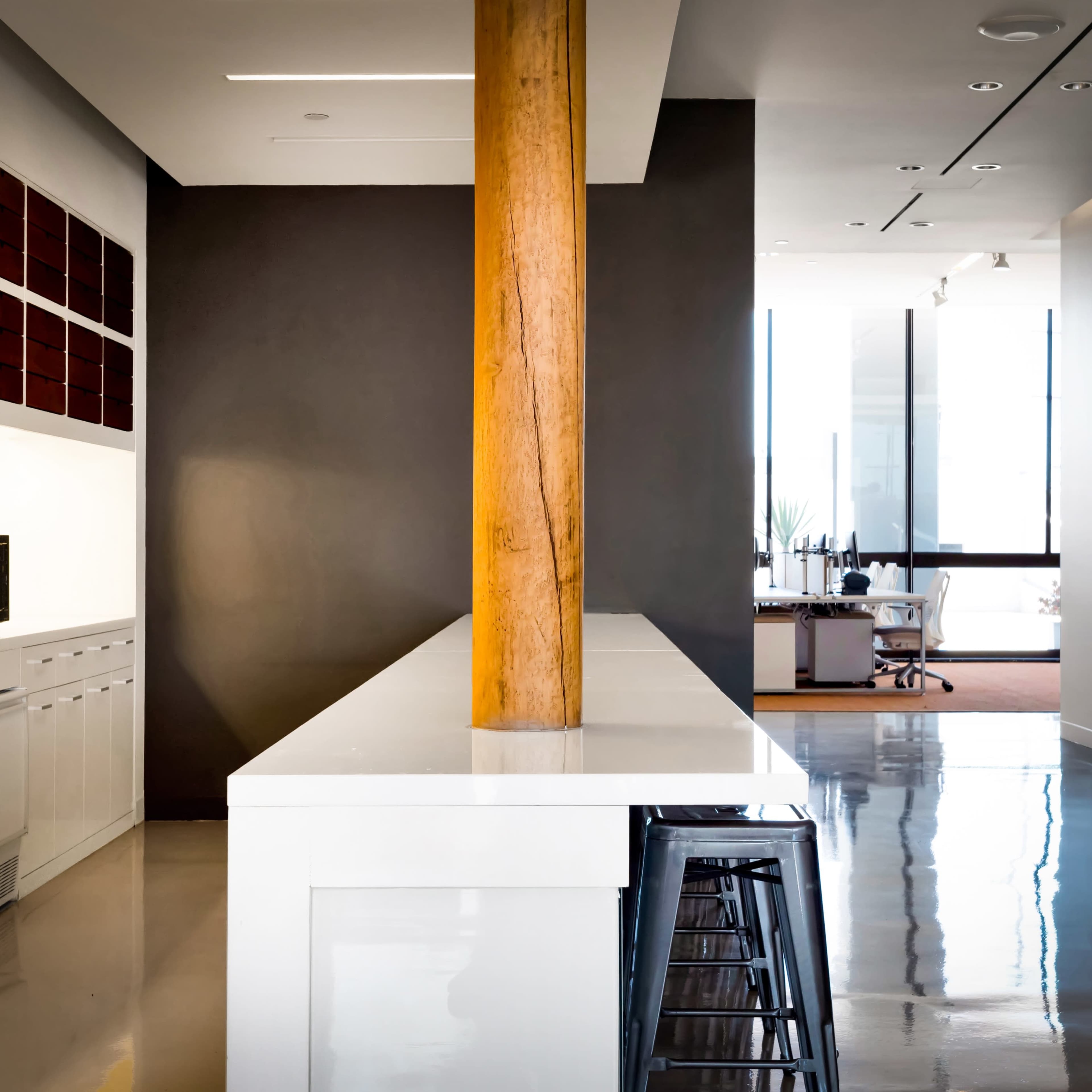 A modern kitchen area with a white countertop, a wooden column, and a workspace in the background featuring a desk and large windows.