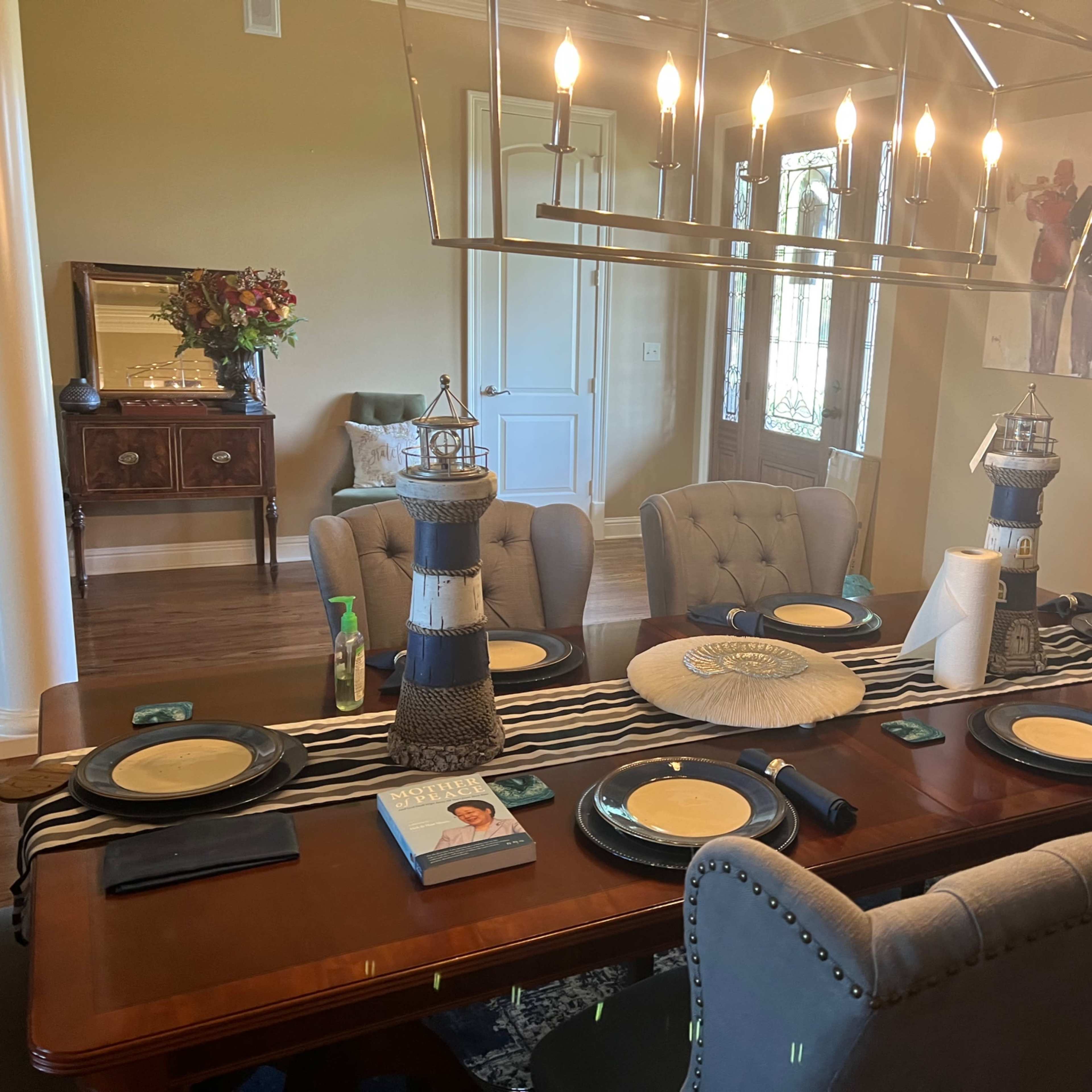 A dining table is set with plates and decorative lanterns, surrounded by upholstered chairs in a room featuring a floral arrangement and artwork on the walls.