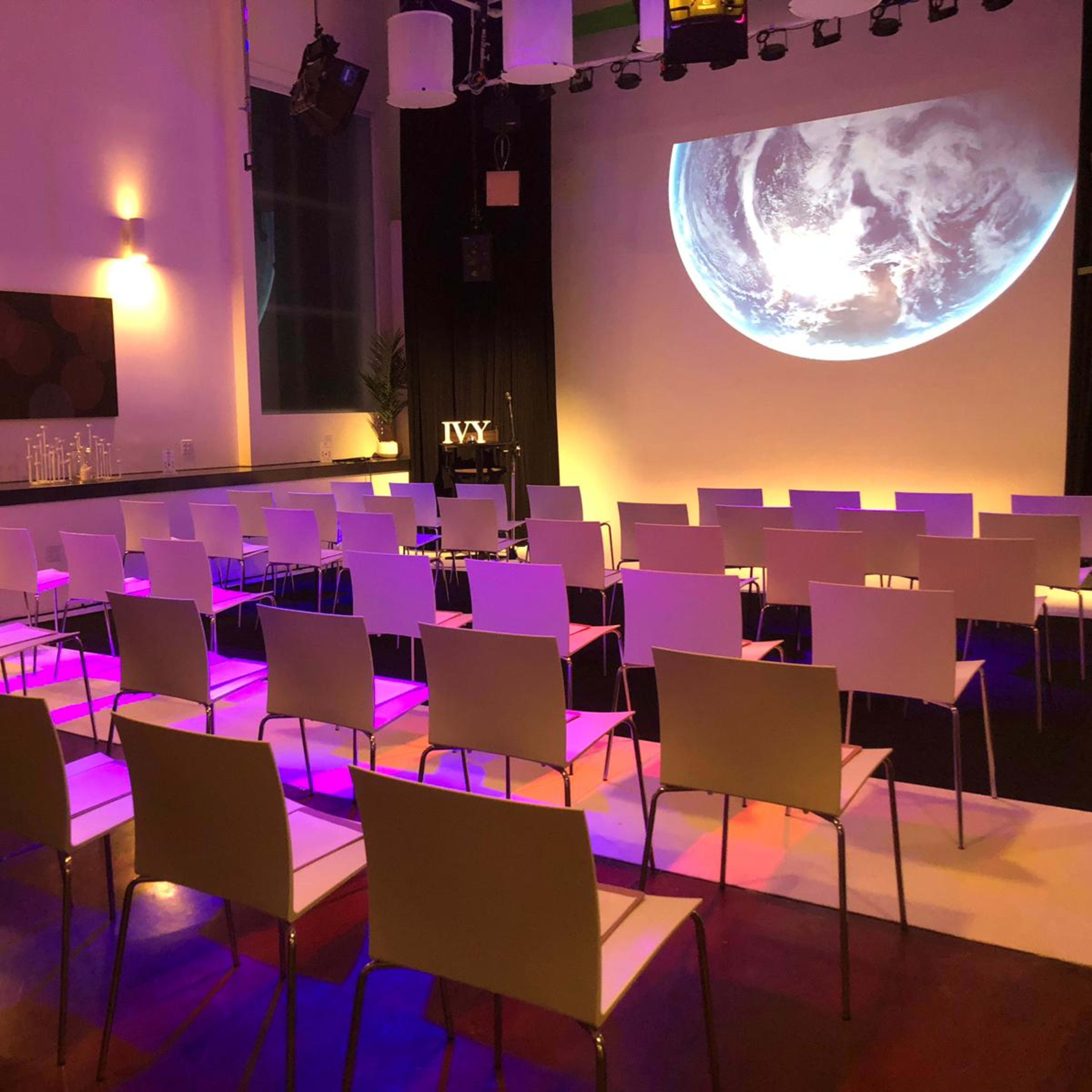 A room is set up for a presentation, featuring rows of white chairs facing a large projection of the Earth on a screen, with colored lighting and a small table in the corner.