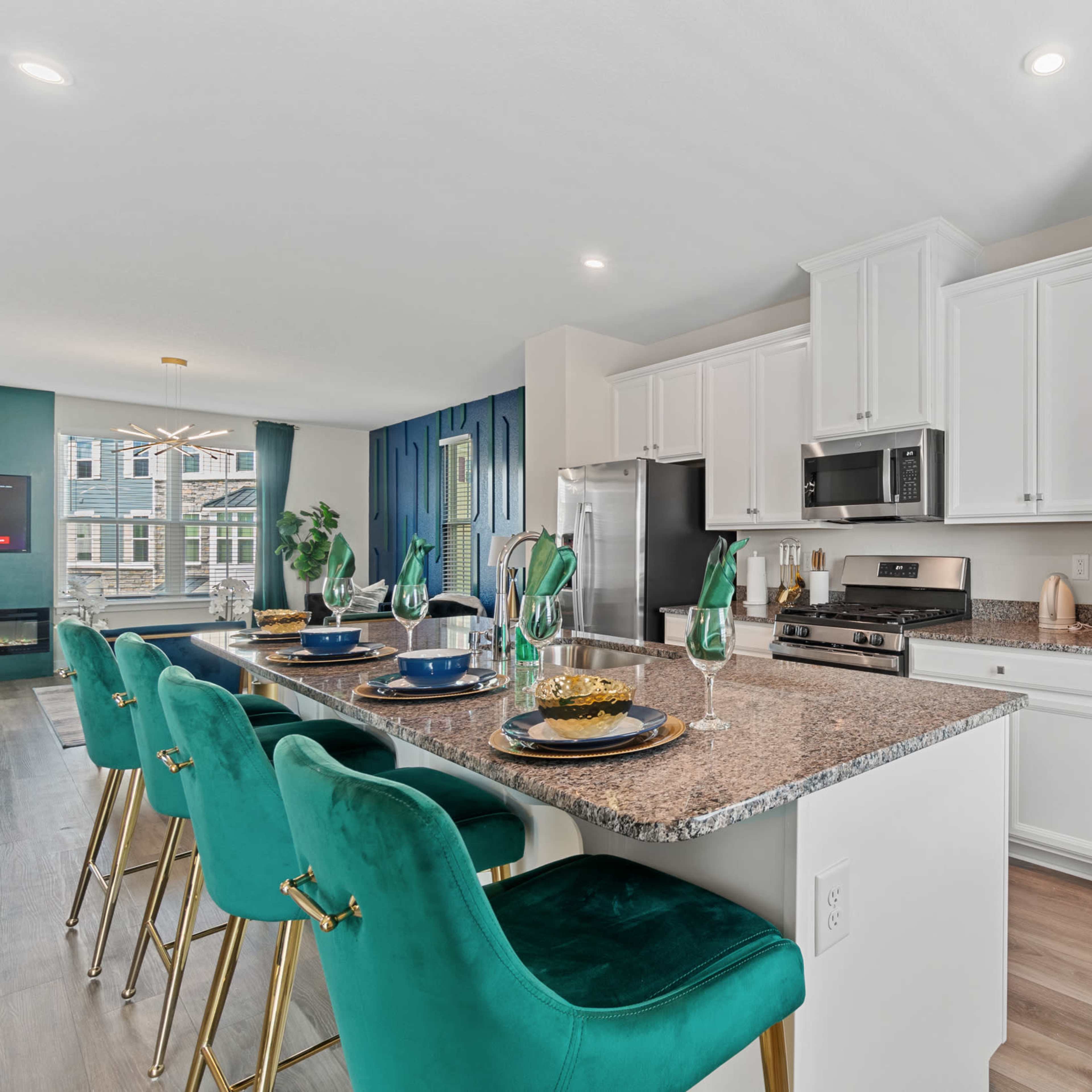 A modern kitchen with a granite countertop, white cabinetry, stainless steel appliances, and green upholstered bar stools.