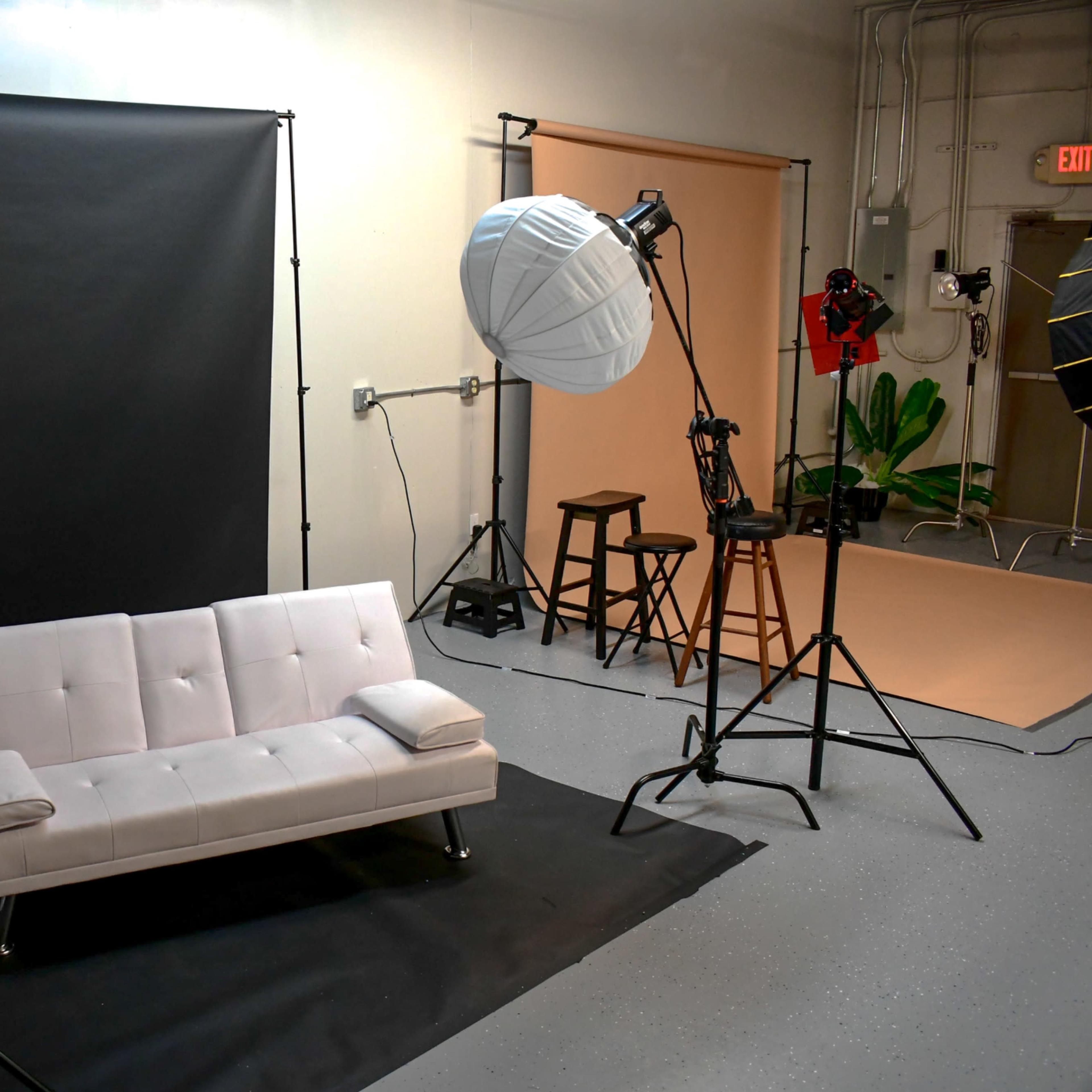 A photography studio setup featuring a white couch on a black backdrop, with various lighting equipment and a stool in the background.